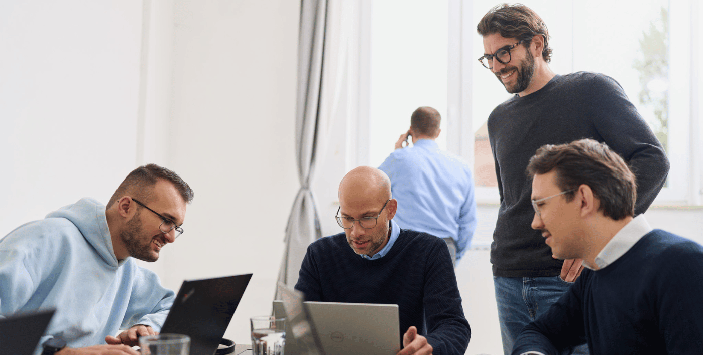 Fünf Männer von loyos bi, drei sitzen am Schreibtisch und schauen auf ihre Laptops, einer steht dahinter und schaut auch darauf, ein anderer telefoniert im Hintergrund.