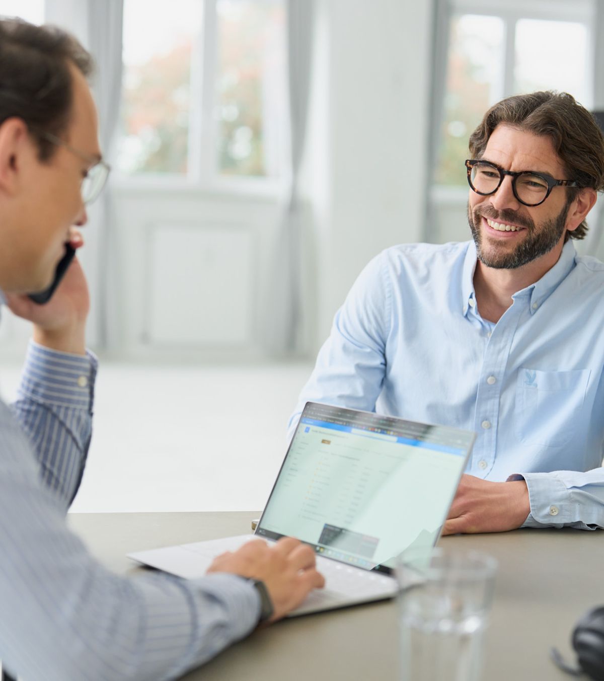 Zwei Mitarbeiter von loyos bi sitzt am Tisch, einer hat seinen Laptop vor sich und ein Handy am Ohr, er telefoniert. Der andere schaut ihr an und lächelt.