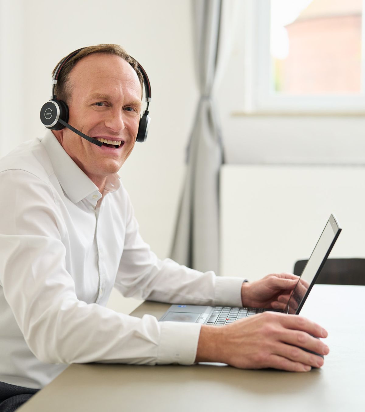 Ein Mann lacht stark, hat ein weißes Hemd an, sitzt am Laptop und hat ein Headset auf. Er arbeitet bei loyos bi.