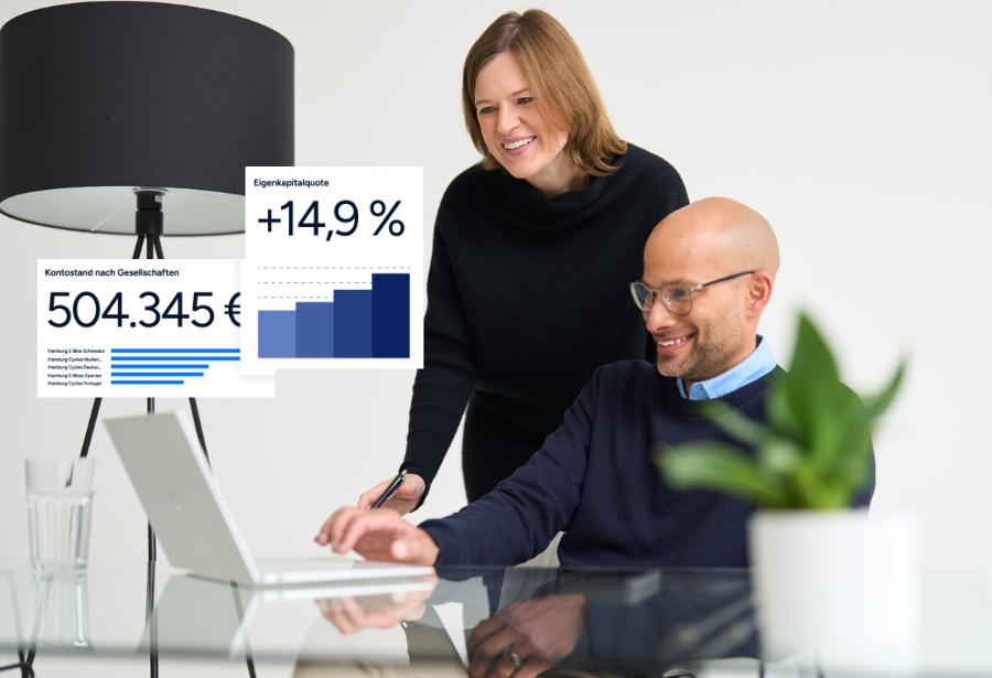 A man is sitting at a table in front of his laptop, a woman is leaning over his shoulder, and they are both laughing.