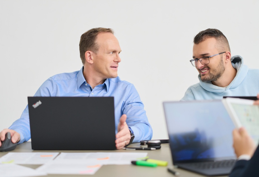 Zwei Männer von loyos bi sitzen am Tisch, ihre Laptops vor sich, sie unterhalten sich, einer lächelt.