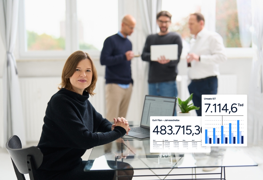 Eine Frau von loyos bi sitzt am Tisch, der Laptop vor sich, dahinter stehen drei Männer, die ins Gespräch vertieft sind.