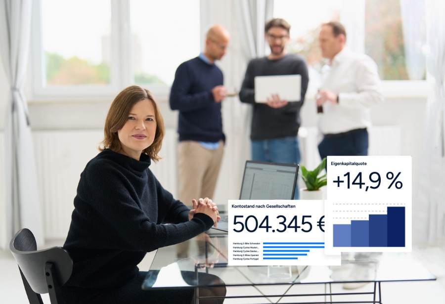 Eine Frau von loyos bi sitzt am Tisch, der Laptop vor sich, dahinter stehen drei Männer, die ins Gespräch vertieft sind.