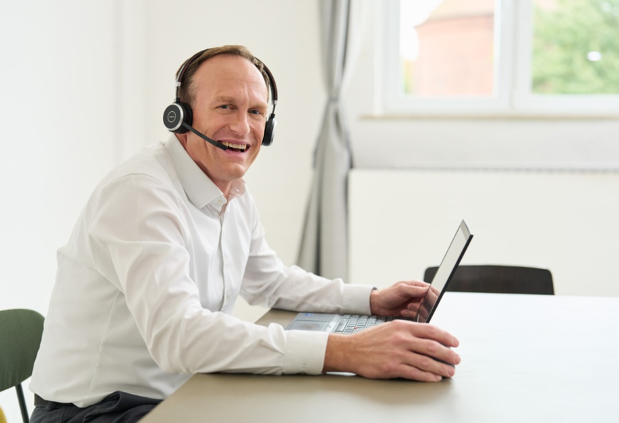 Ein Kollege von loyos bi hat ein Headset auf, sein Laptop vor ihm, er sitzt an einem großen Tisch, lächelt in die Kamera.