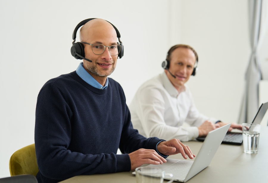 Zwei Kollegen mit Headset und Laptop sitzen am Tisch, beide lächeln in die Kamera.