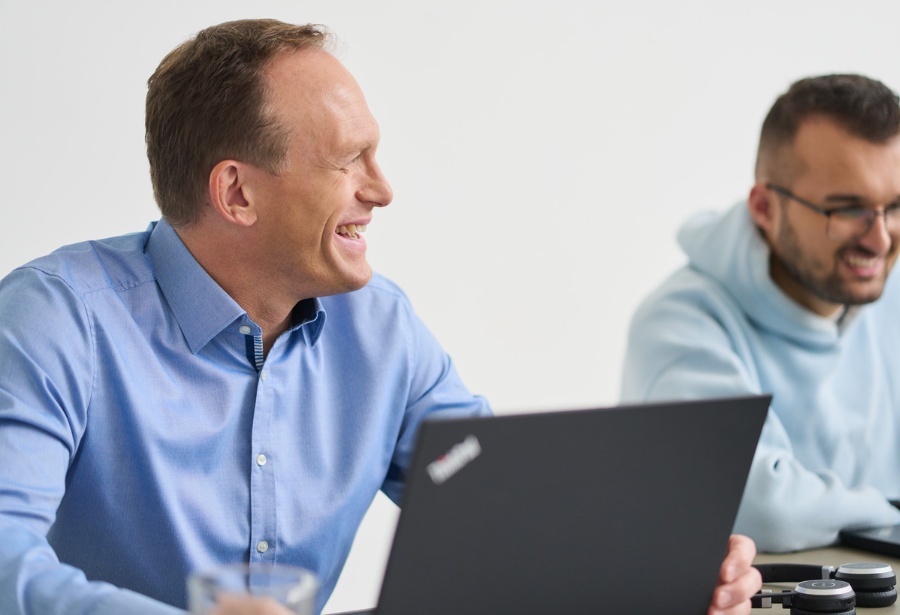 Ein Kollege von loyos bi sitzt am Laptop, schaut seinen Kollegen an und lacht.