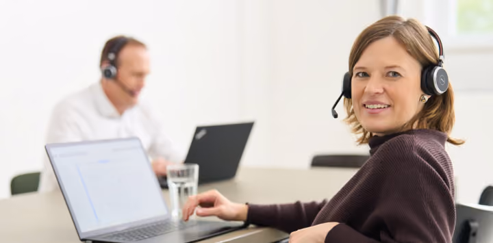 Frau mit Headset arbeitet an Laptop, im Hintergrund Mann mit Headset am Laptop in Büro.