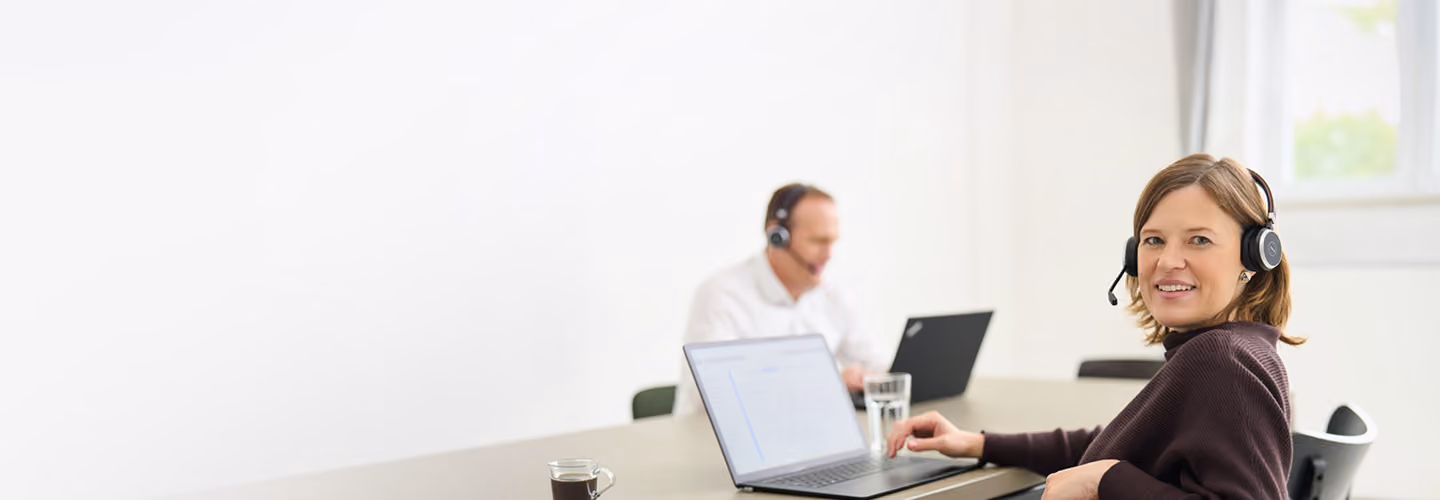 Zwei Personen mit Headsets sitzen an einem Tisch und arbeiten an Laptops in einem hellen Büro.