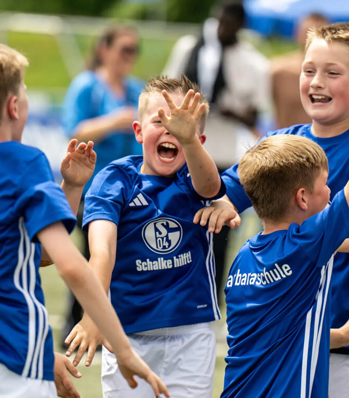Young boys in blue soccer jerseys celebrating energetically on a field, with one boy raising his hand and smiling widely.