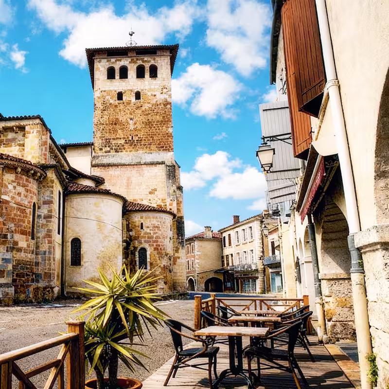 Vue contemporaine de la place et de l’église avec terrasse du Bistrot des Arceaux sous un ciel bleu.