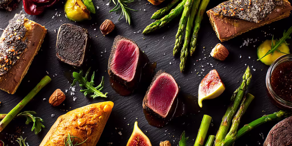 Plateau gastronomique avec tranches de magret saignant, foie gras, asperges vertes, figues fraîches et éclats de noisettes sur ardoise.