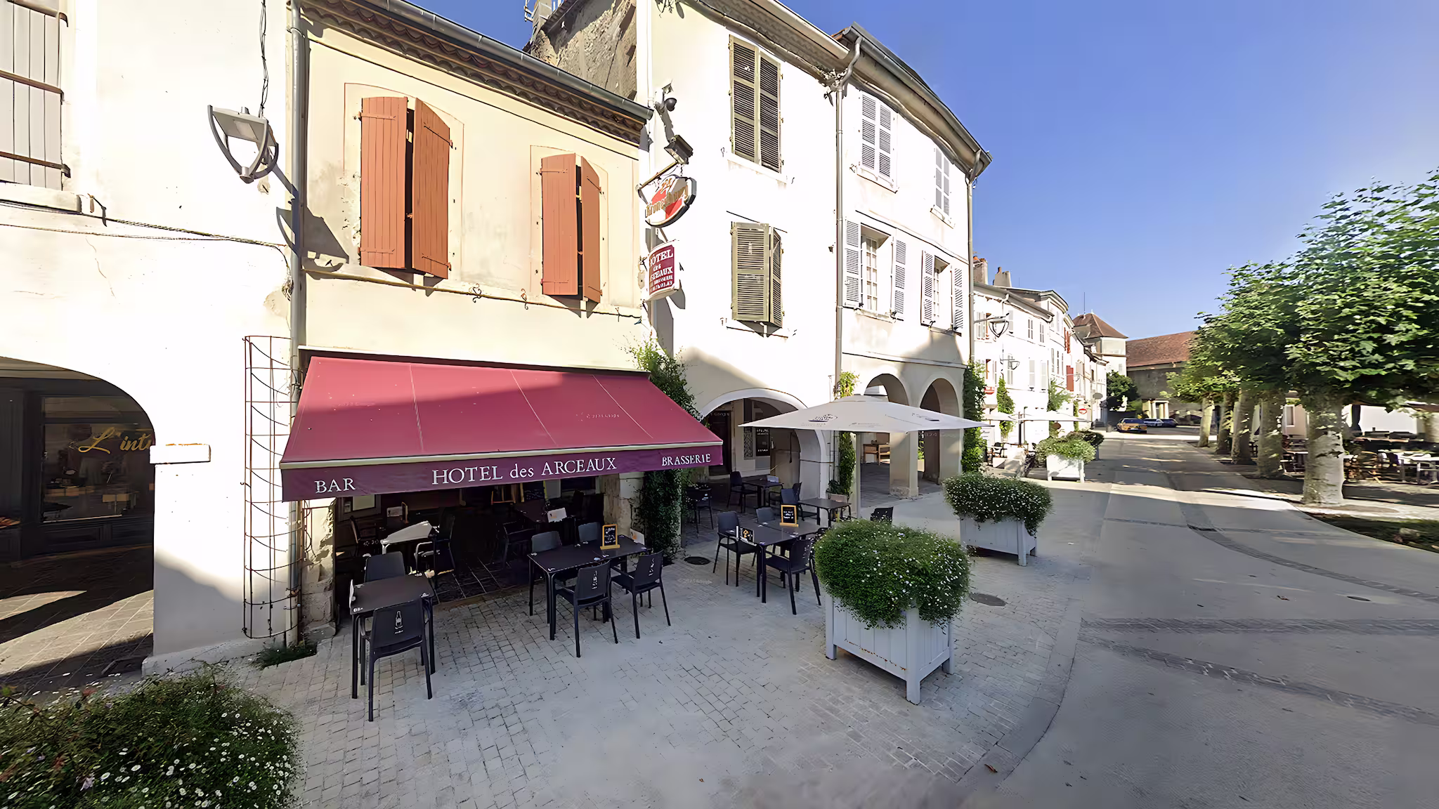 Façade claire de l’Hôtel des Arceaux à Saint-Sever, terrasse ombragée et rue bordée de platanes sous un ciel bleu d’été.