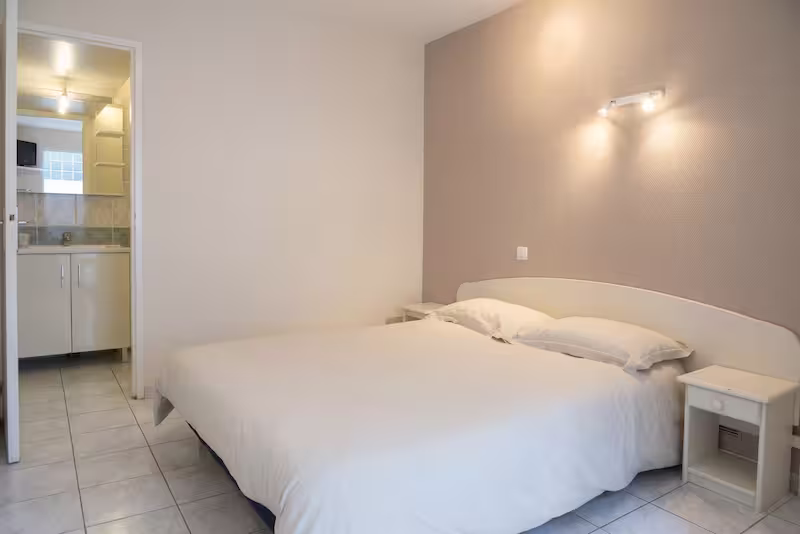 Chambre double avec un grand lit blanc, deux tables de chevet et une porte donnant sur une petite salle de bain privative.