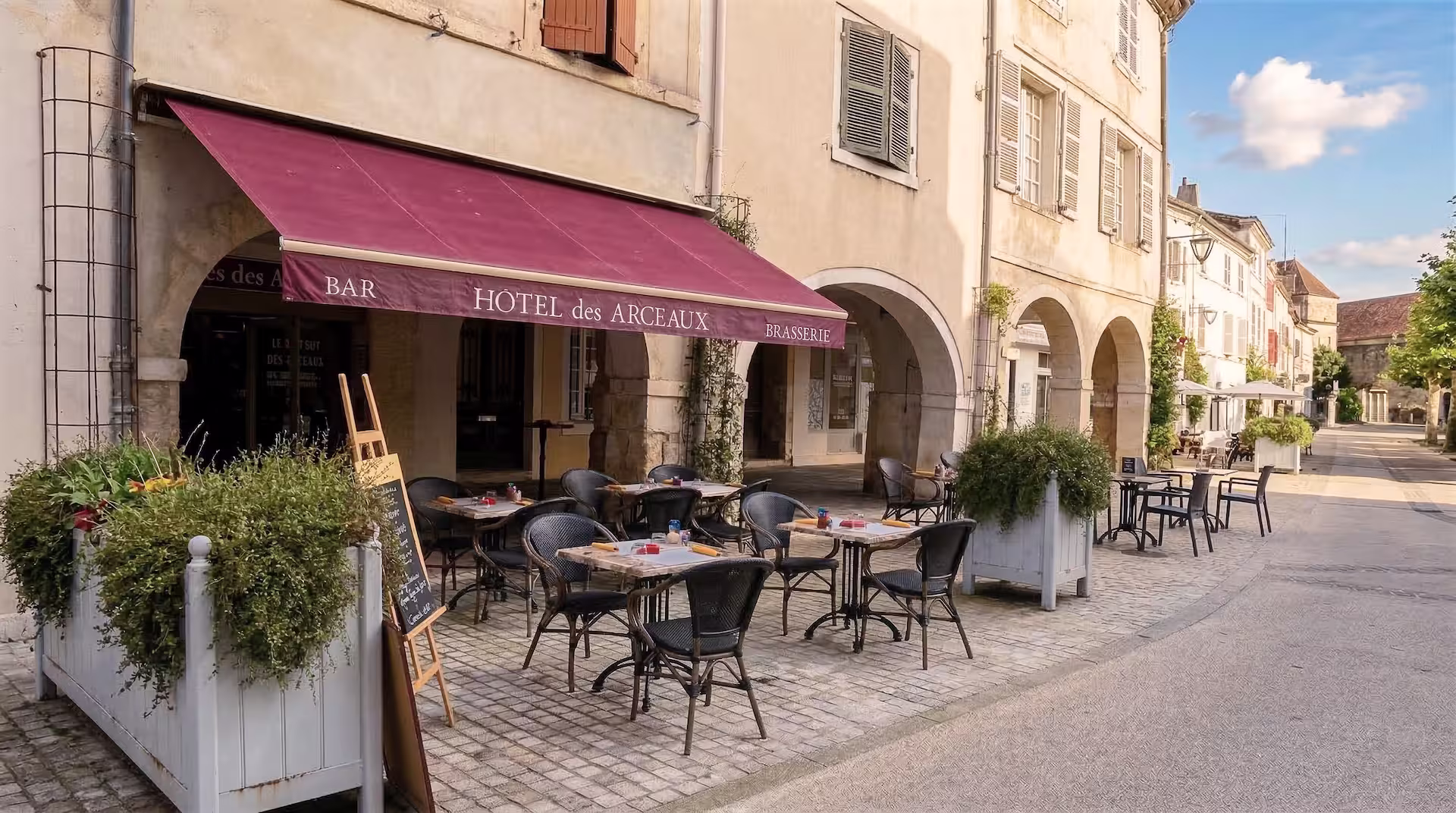 Terrasse extérieure d'un bar-brasserie avec tables et chaises en rotin sous un auvent bordeaux marqué Hôtel des Arceaux.