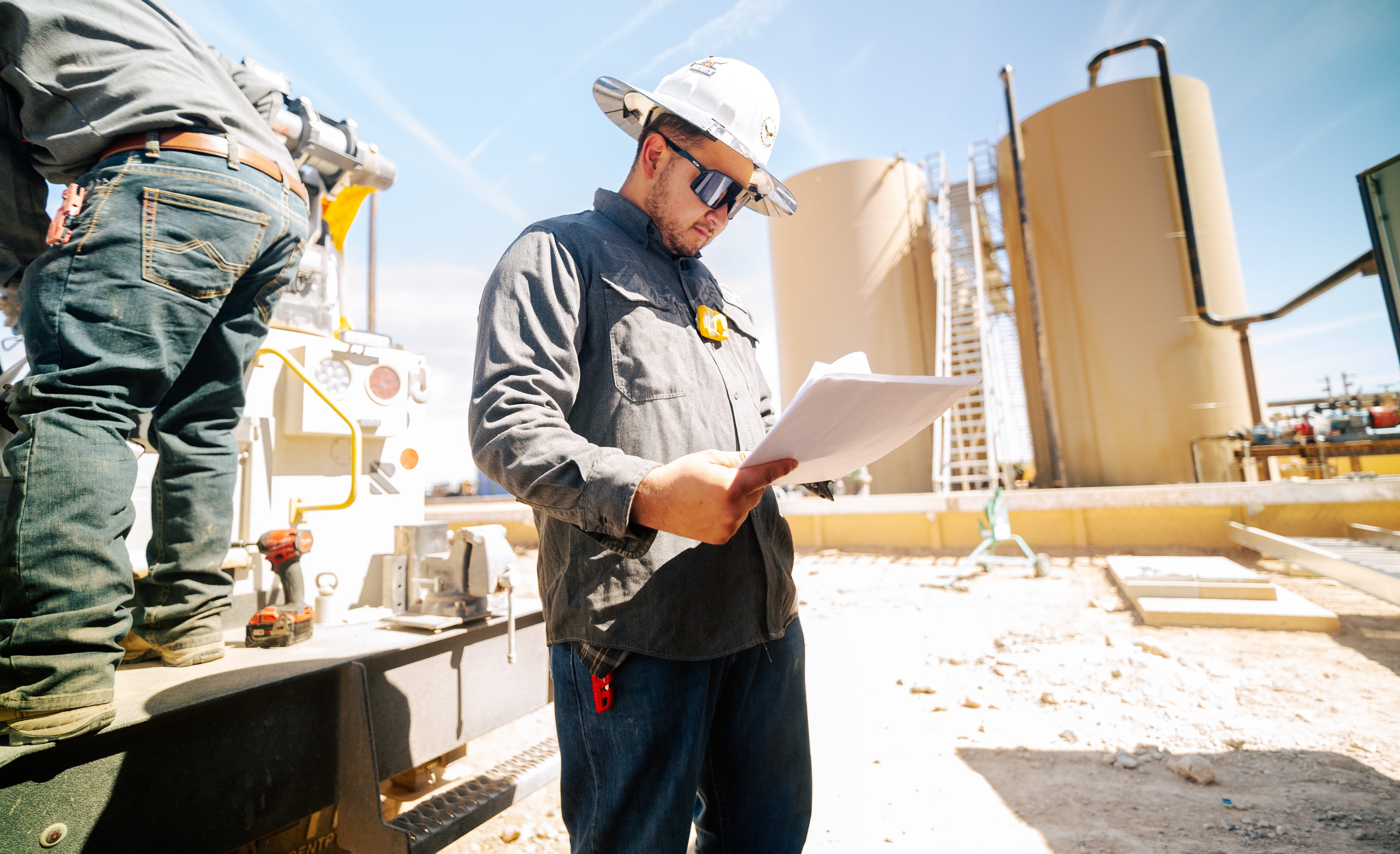 Construction worker wearing a white hard hat and sunglasses reads documents at an industrial site with large storage tanks in the background on a sunny day.