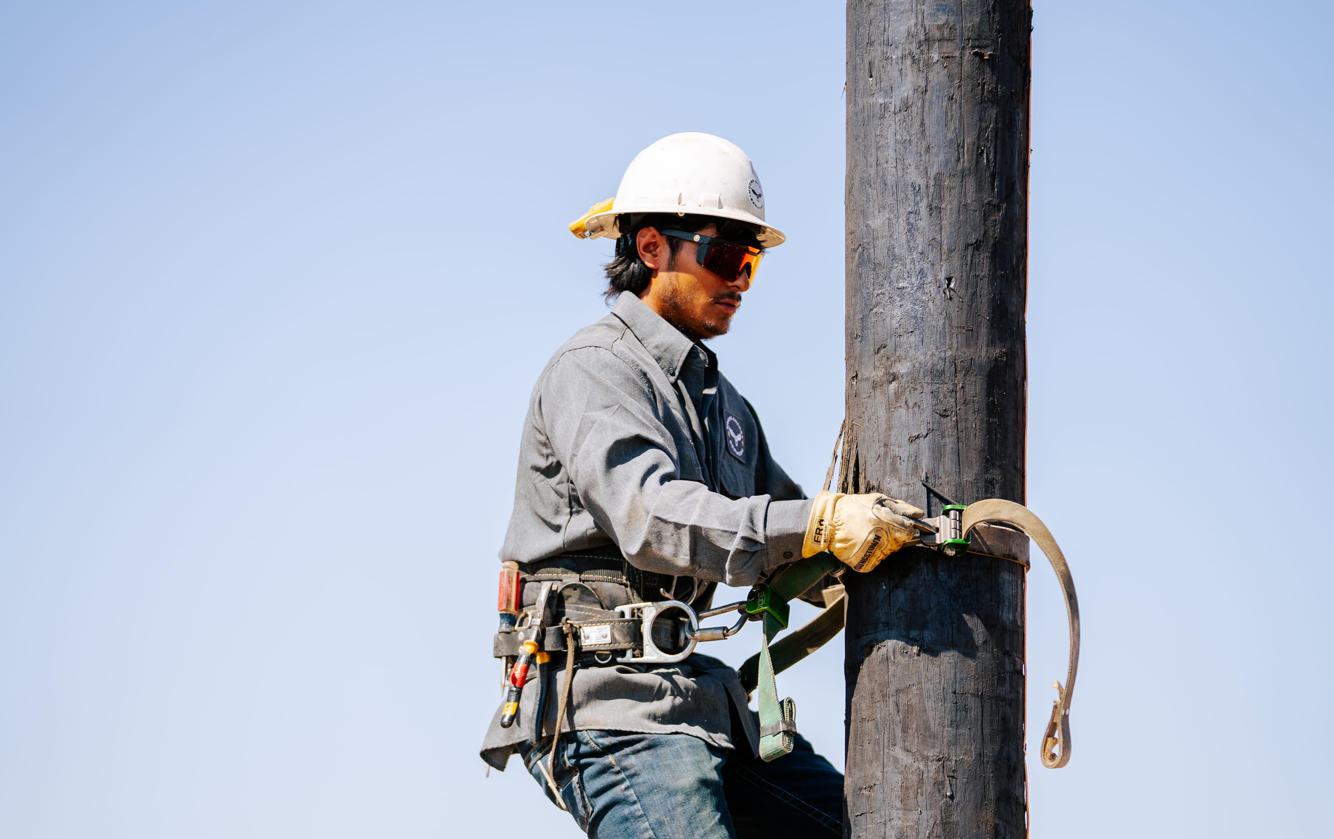 Utility worker wearing a white hard hat, safety glasses, and gloves is securing a safety harness strap around a wooden utility pole.