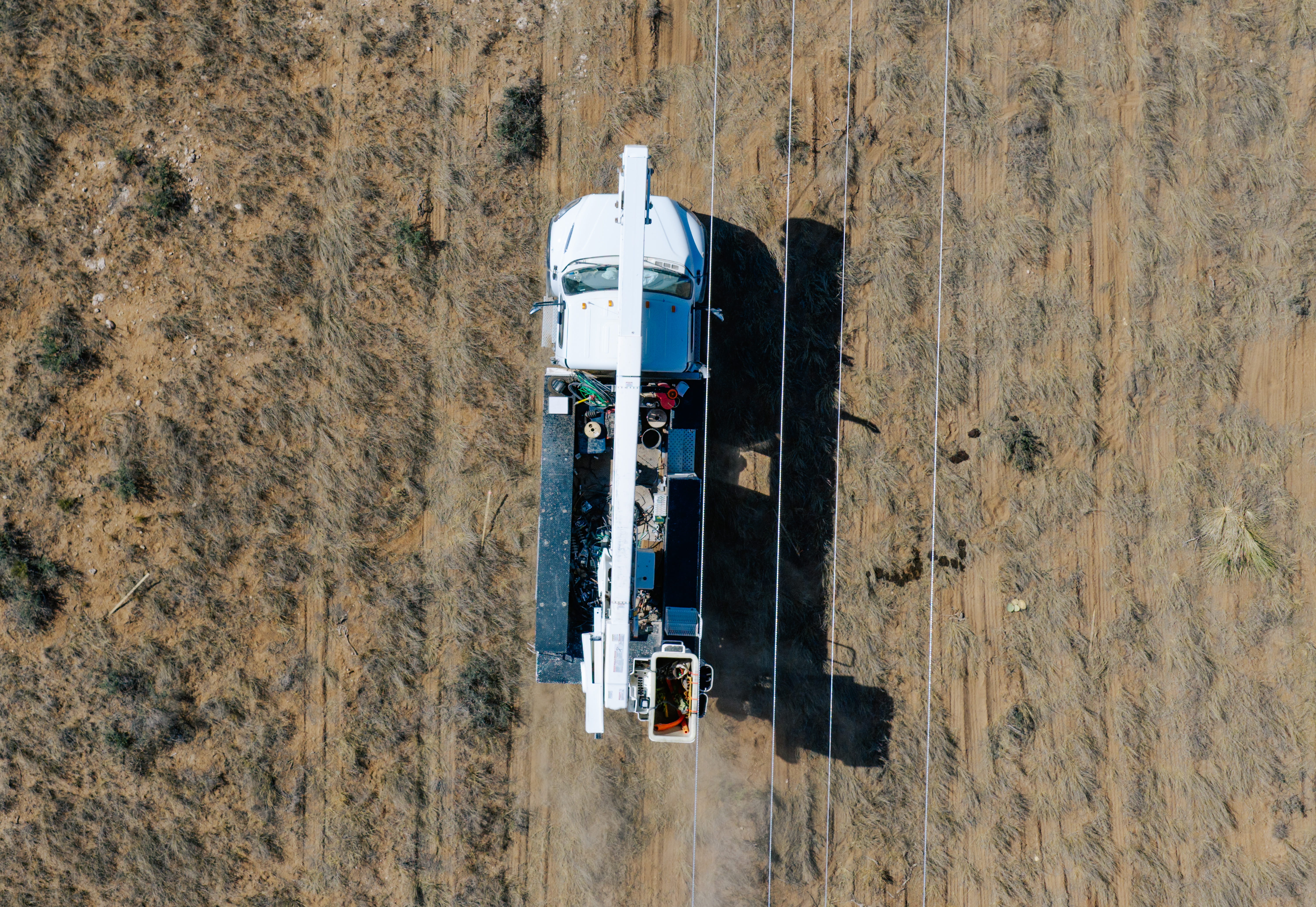 Top-down view of a white utility truck with an extended boom lift near overhead power lines on dry, sparsely vegetated ground.