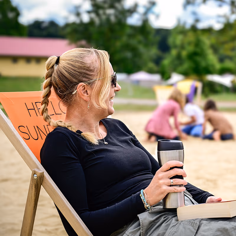 Eine Frau entspannt im Liegestuhl am Sandstrand, Kinder spielen im Hintergrund.