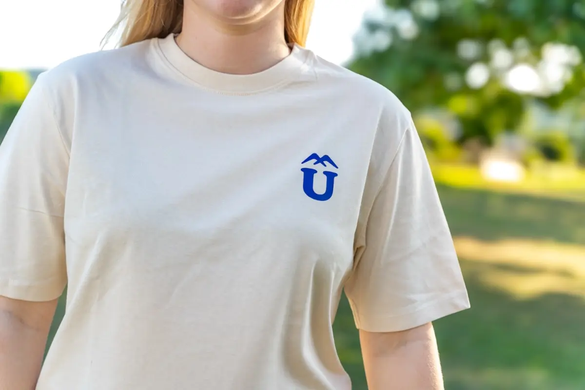 Person trägt beiges T-Shirt mit blauem Überland Logo vor grünem Hintergrund.