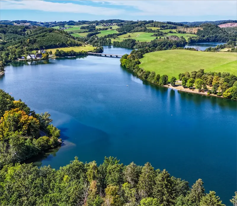 Landschaft mit blauem See, grünen Feldern, Bäumen und einer kleinen Brücke im Hintergrund.