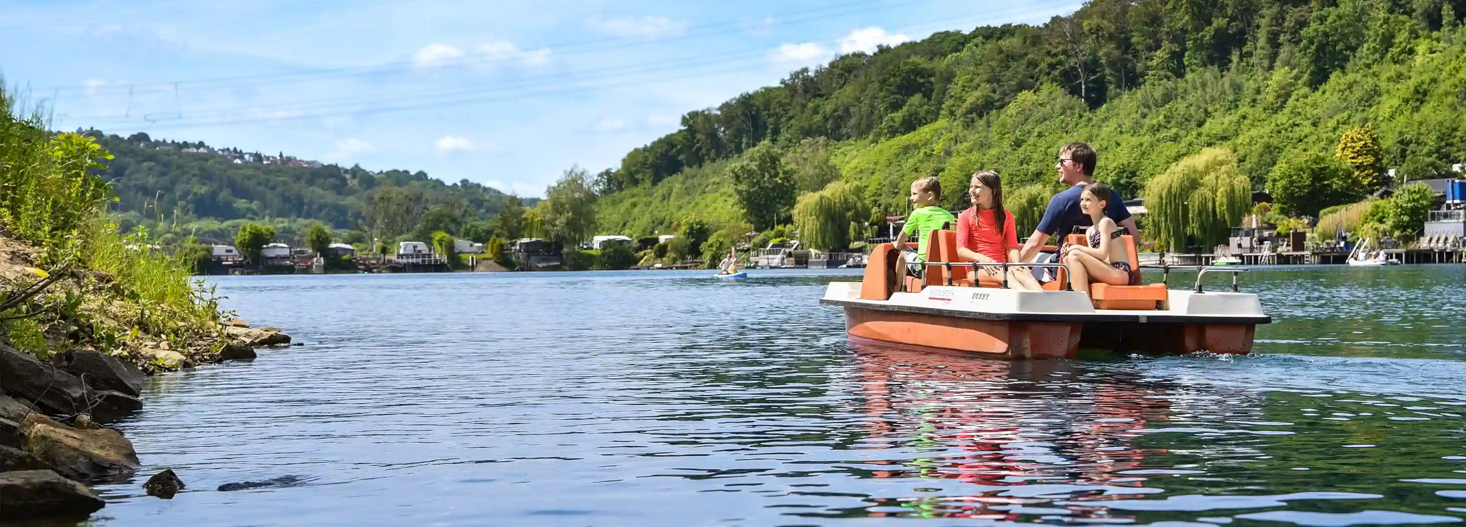 Familie mit zwei Kindern fährt Tretboot auf ruhigem See an bewaldeten Ufern bei sonnigem Wetter.
