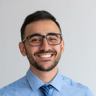 Smiling man with glasses, dark hair, beard, blue shirt, and striped tie on gray background.