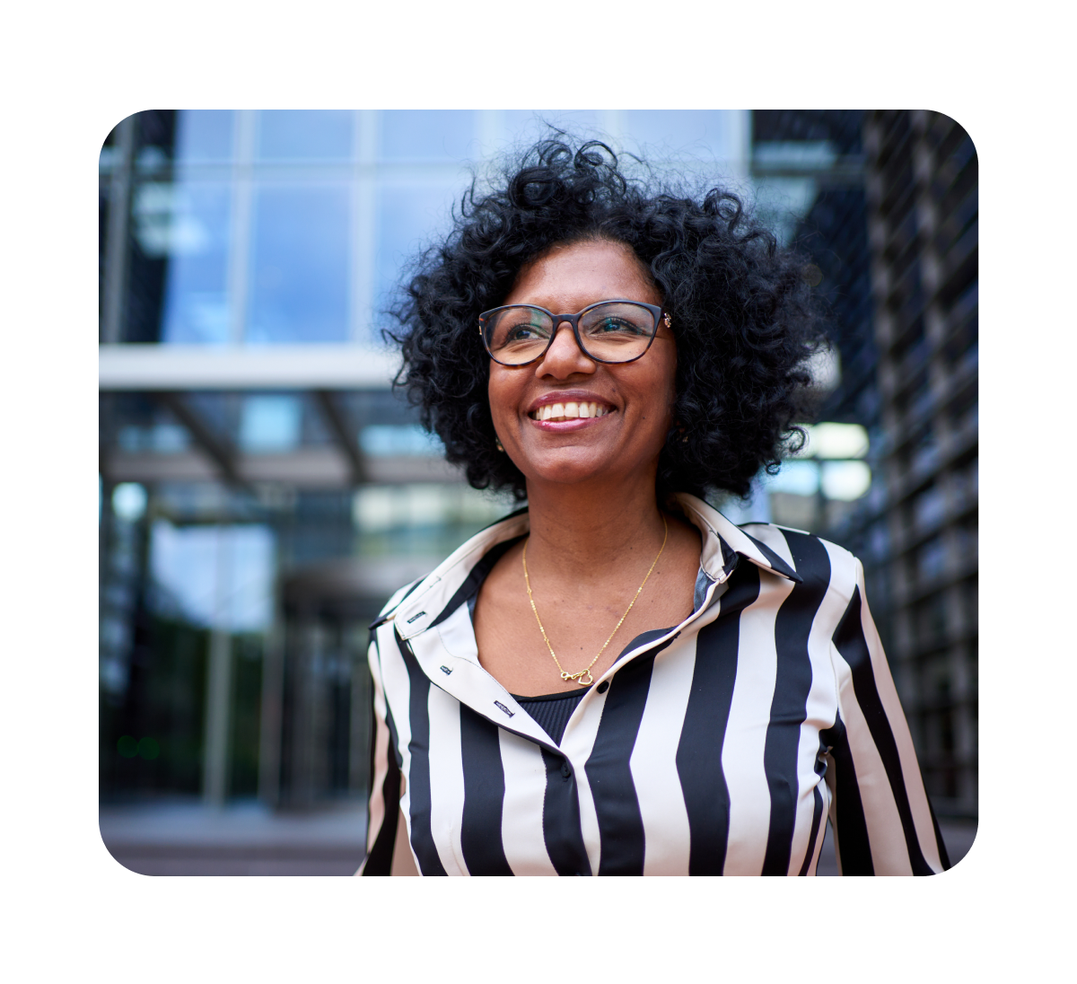 Smiling woman with curly hair and glasses wearing a black and white striped shirt outdoors in front of a modern glass building.