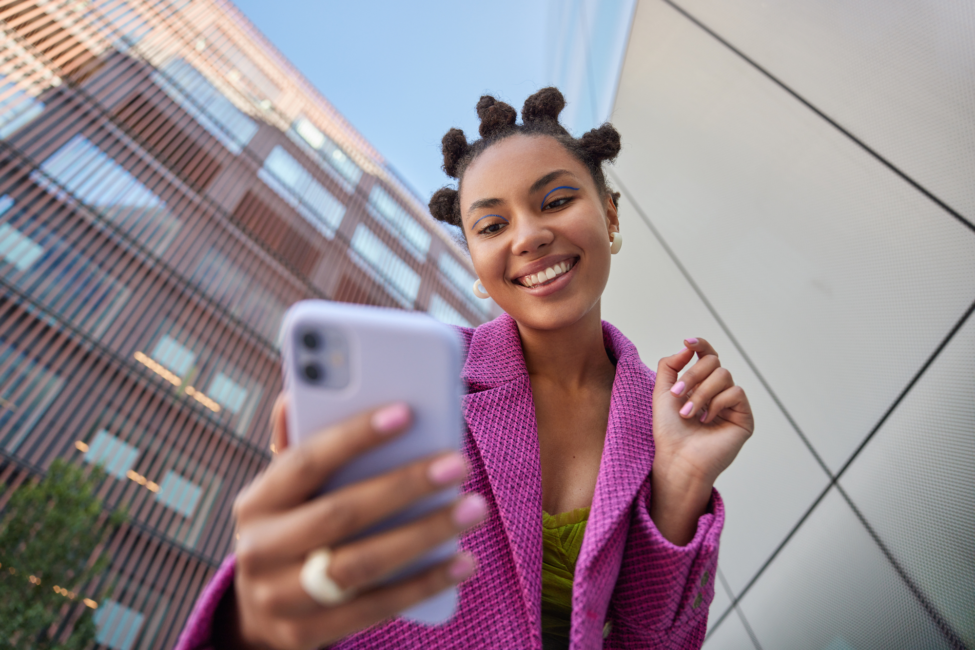Smiling young woman with bantu knot hairstyle wearing a purple blazer taking a selfie on her smartphone outdoors with buildings in the background.