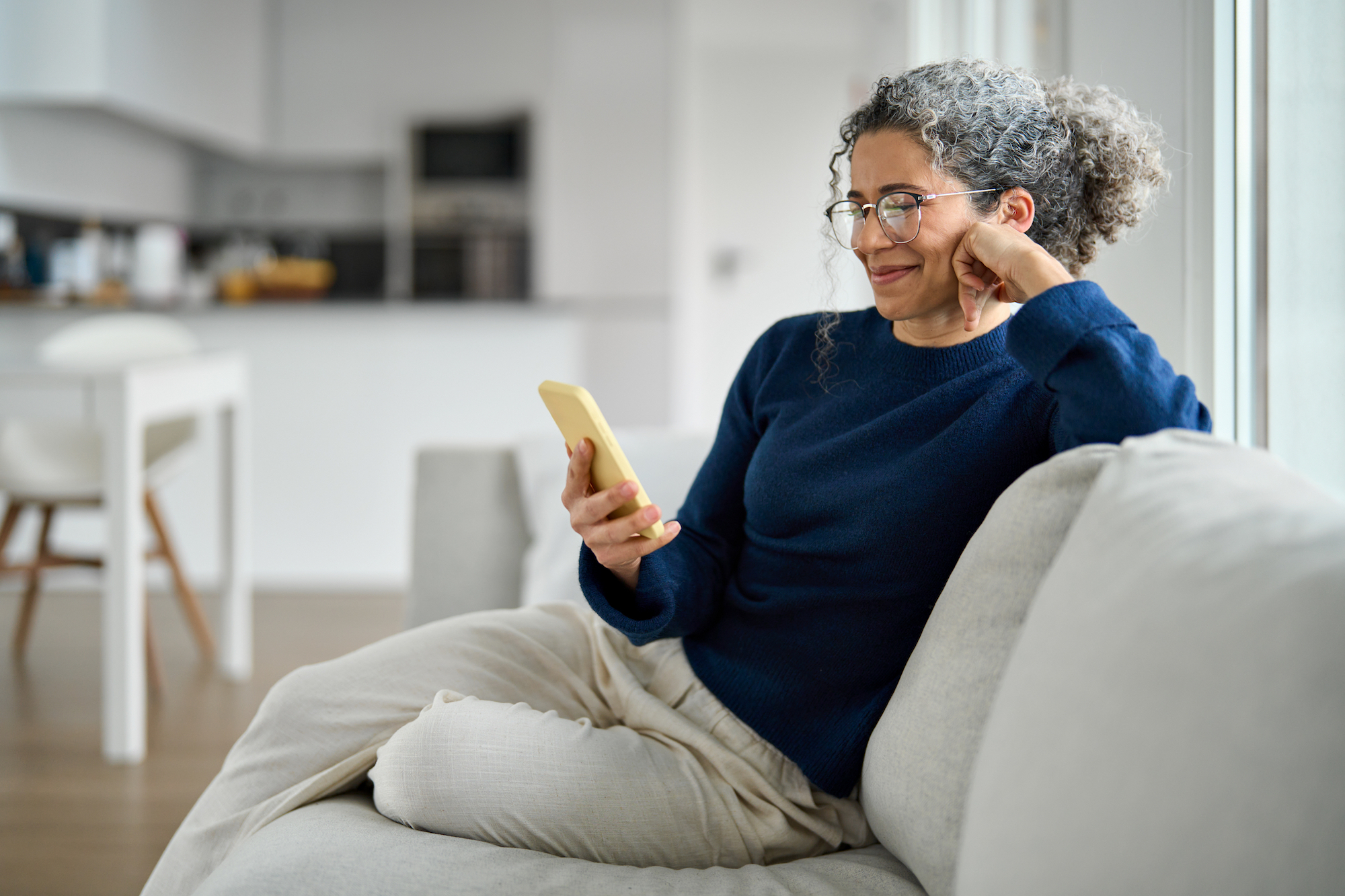 Woman with curly hair and glasses sitting on a couch, smiling while looking at her phone.