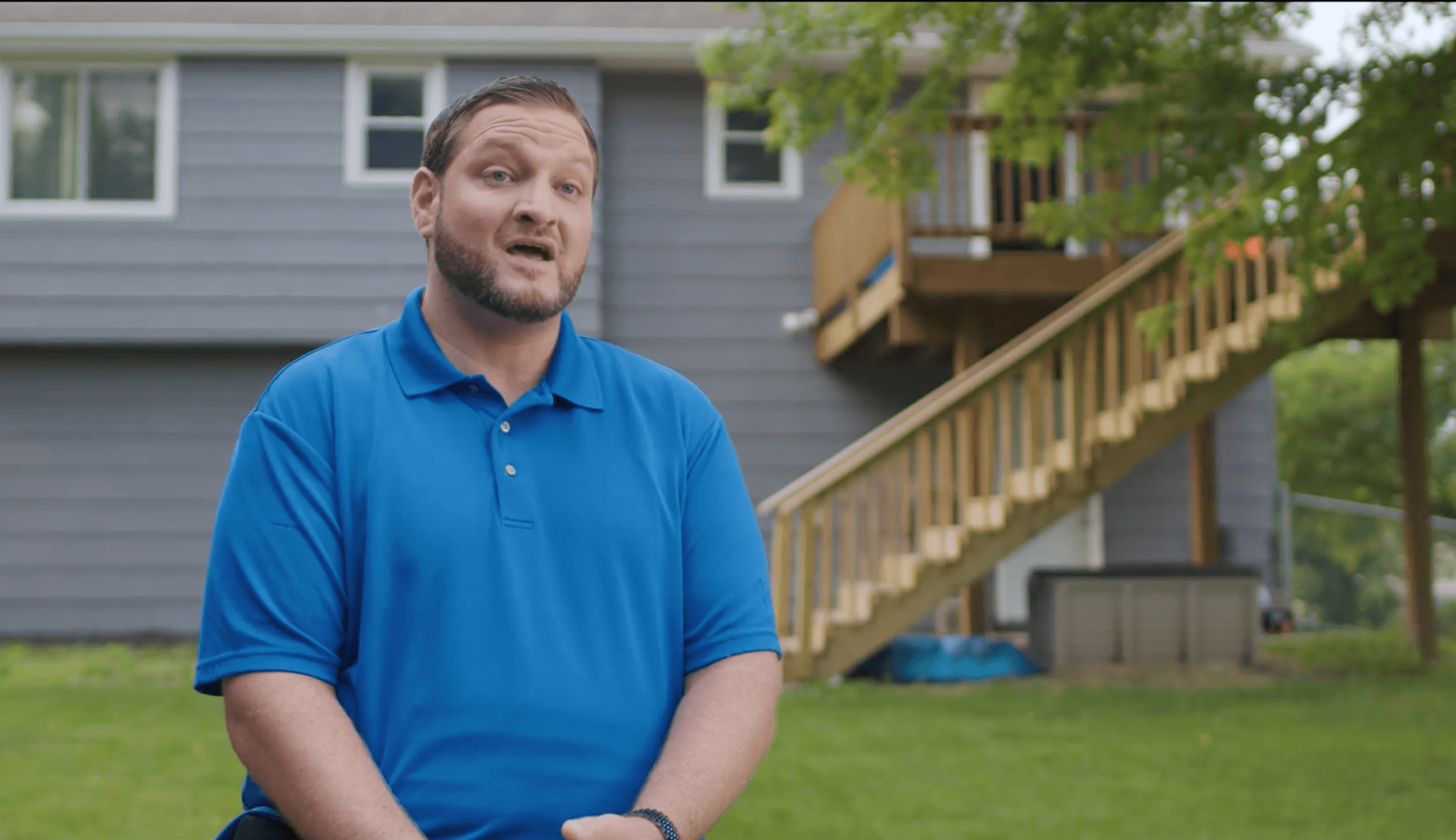 Man in a blue polo shirt speaking with a house and wooden staircase in the background.