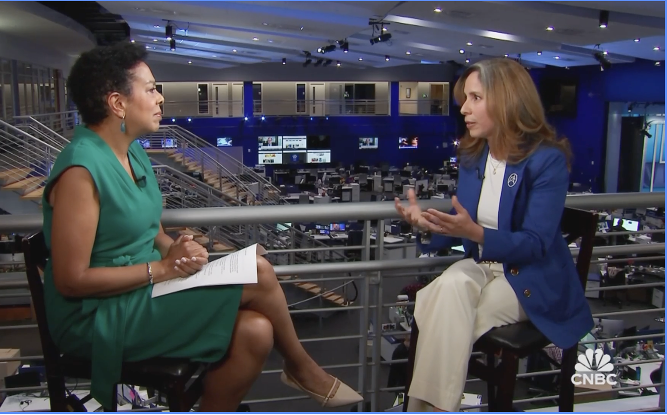 Two women engaged in a conversation while seated in a newsroom with multiple monitors and desks in the background.