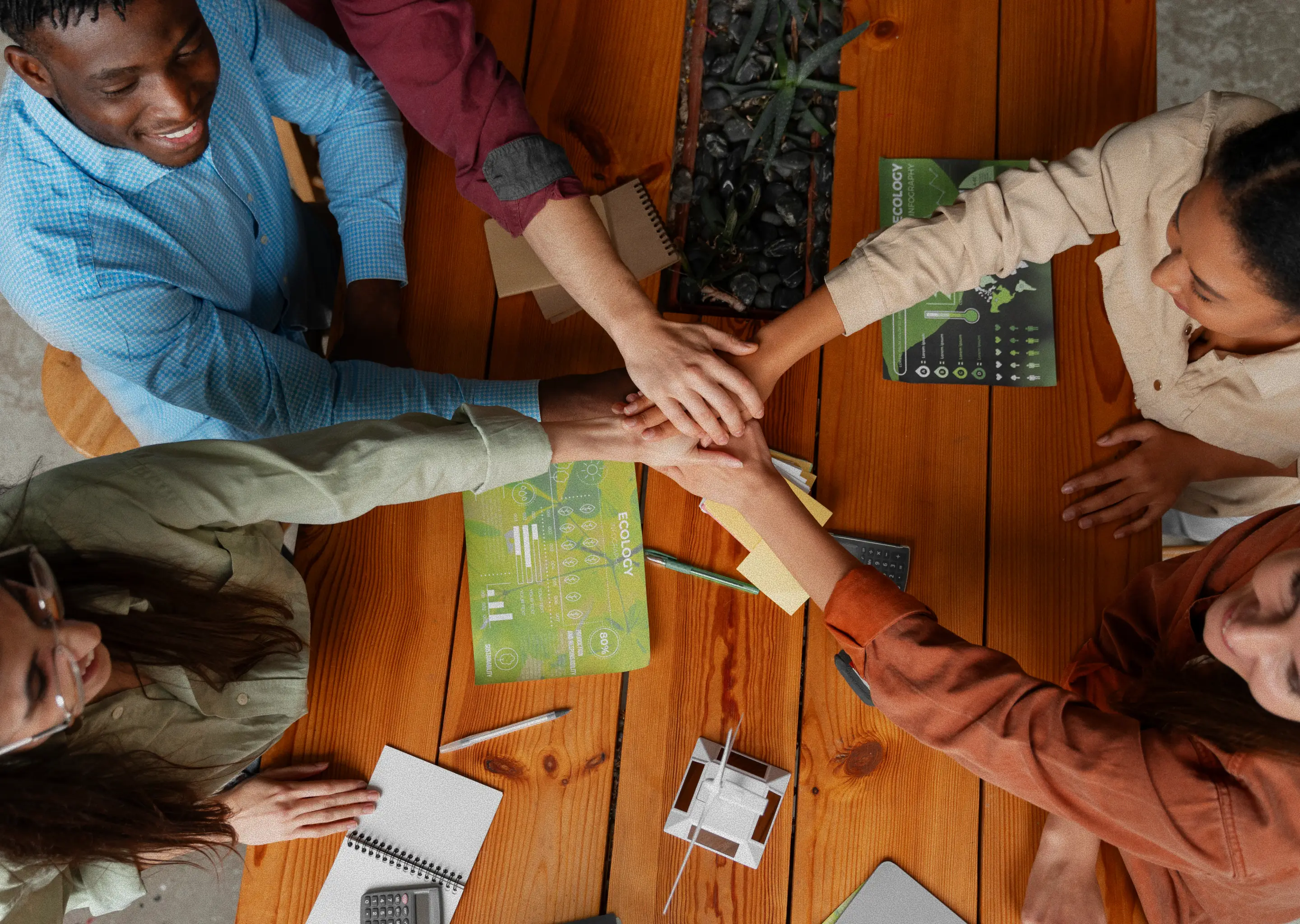 An overhead shot of five smiling coworkers, two men and three women, stacking their hands in the middle of a wooden table in an office.