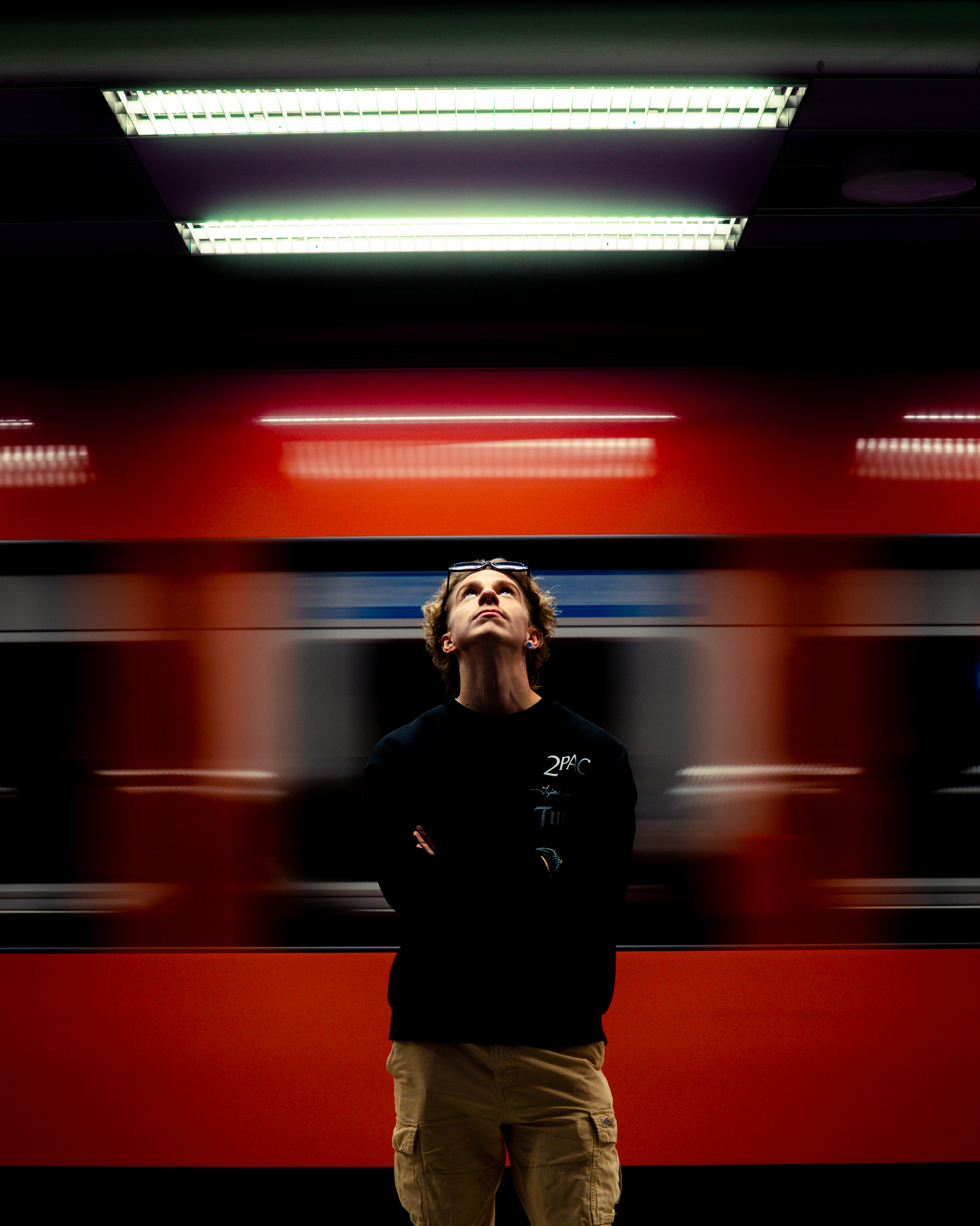 Person in black sweatshirt and tan pants standing in front of a blurred red train, looking up.