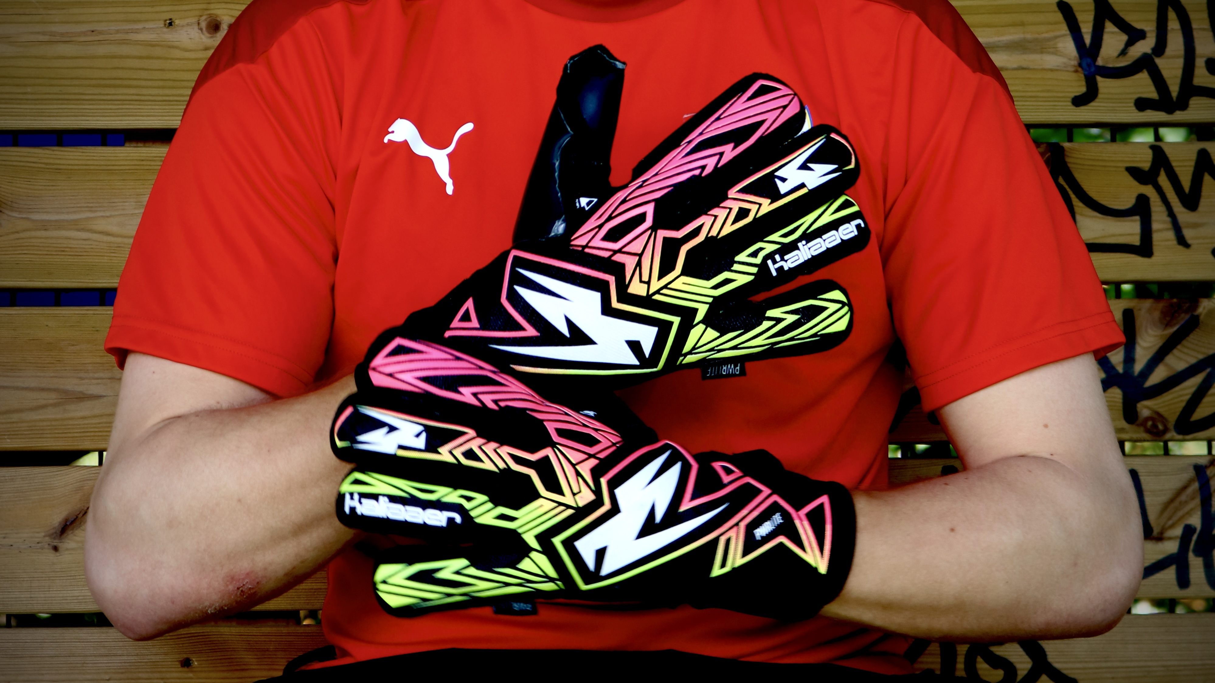 Person wearing red Puma shirt holding up colorful goalkeeper gloves with neon pink, yellow, and black patterns.