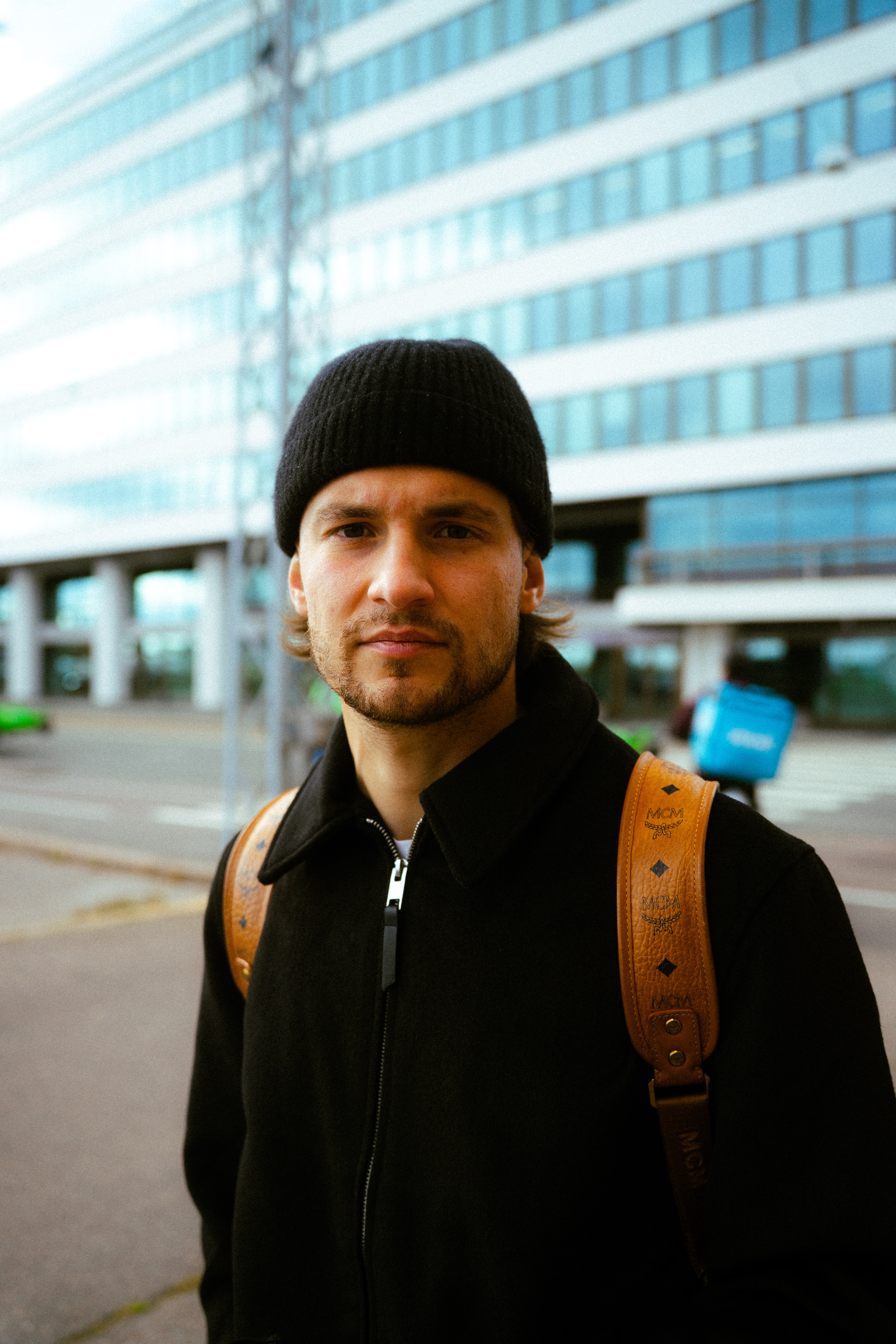 Young man wearing a black beanie and coat with a brown MCM backpack standing in an urban area with a glass building in the background.