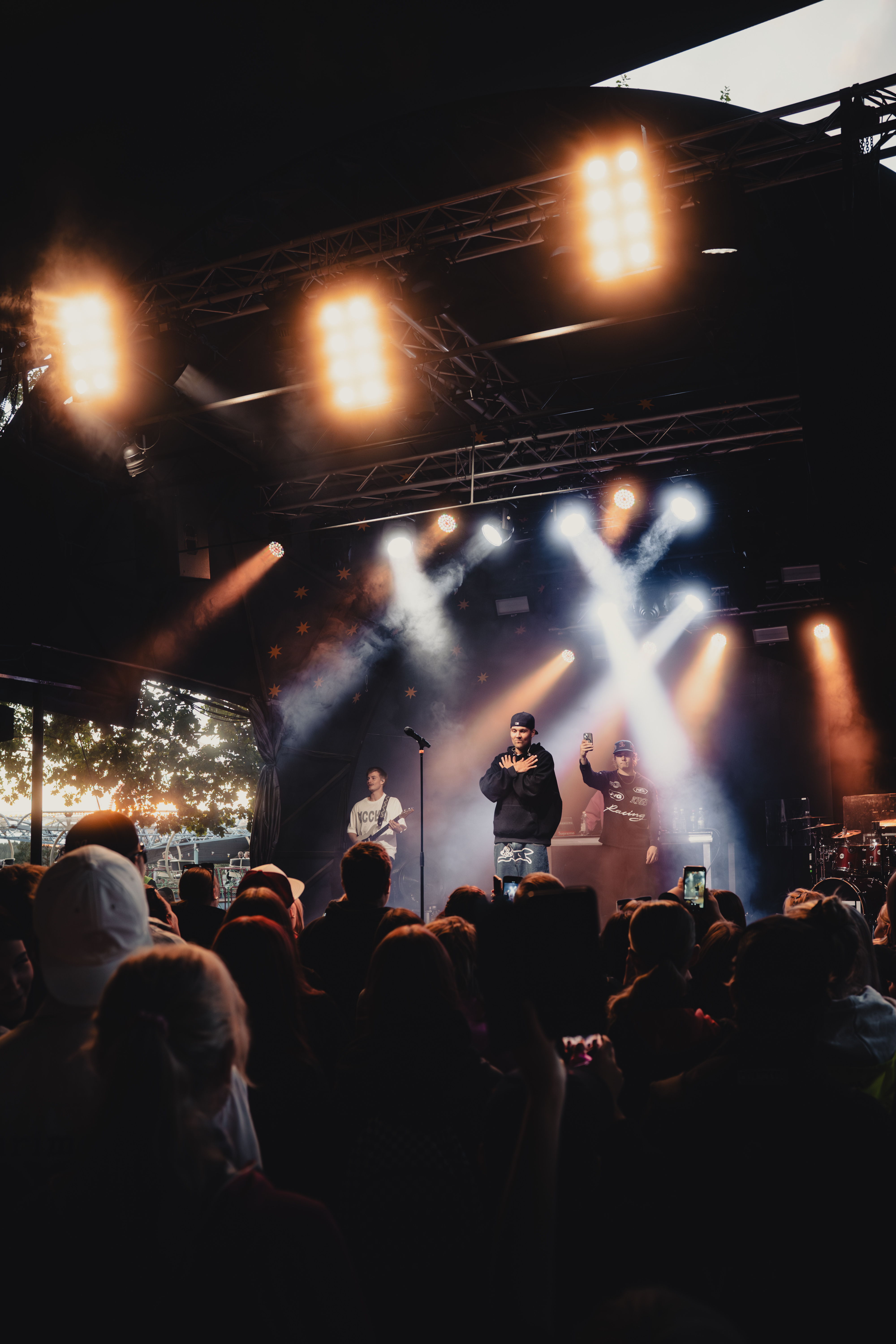 Musicians performing on stage with bright spotlights and an audience watching at an outdoor concert during dusk.
