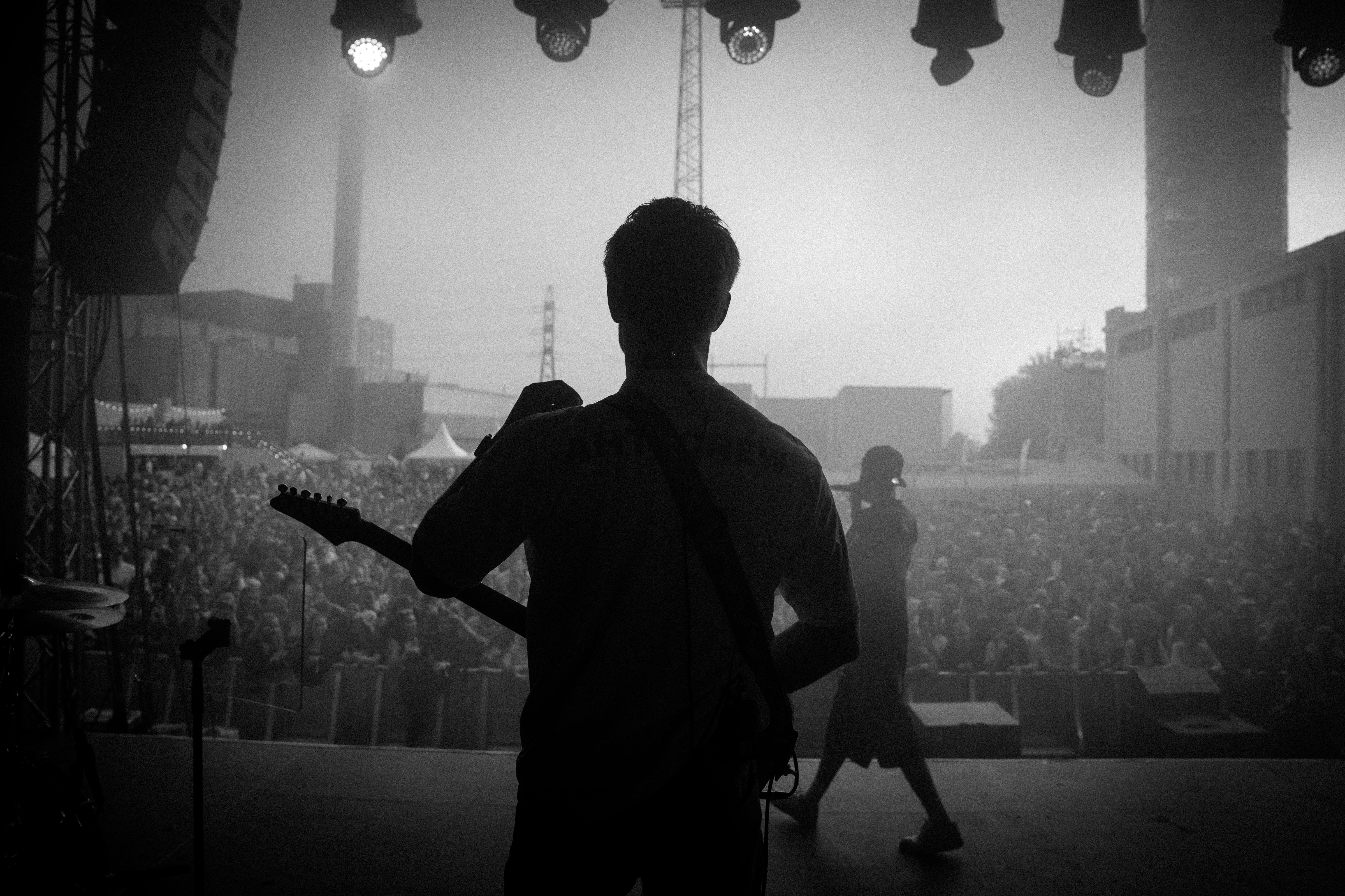 Silhouettes of two musicians on stage facing a large outdoor crowd during a concert at dusk.
