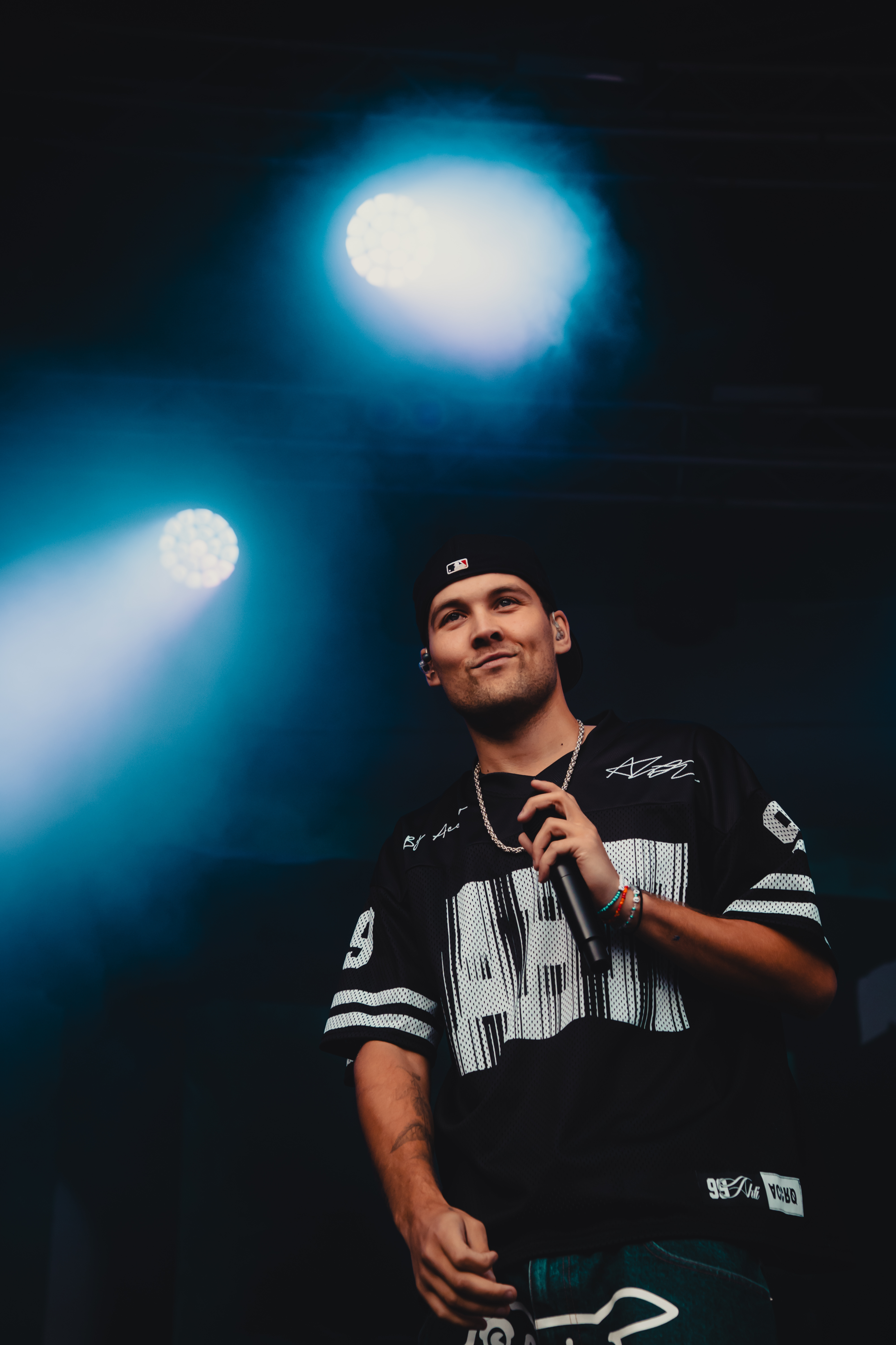 Male performer in black jersey and backward cap holding a microphone under blue stage lights.