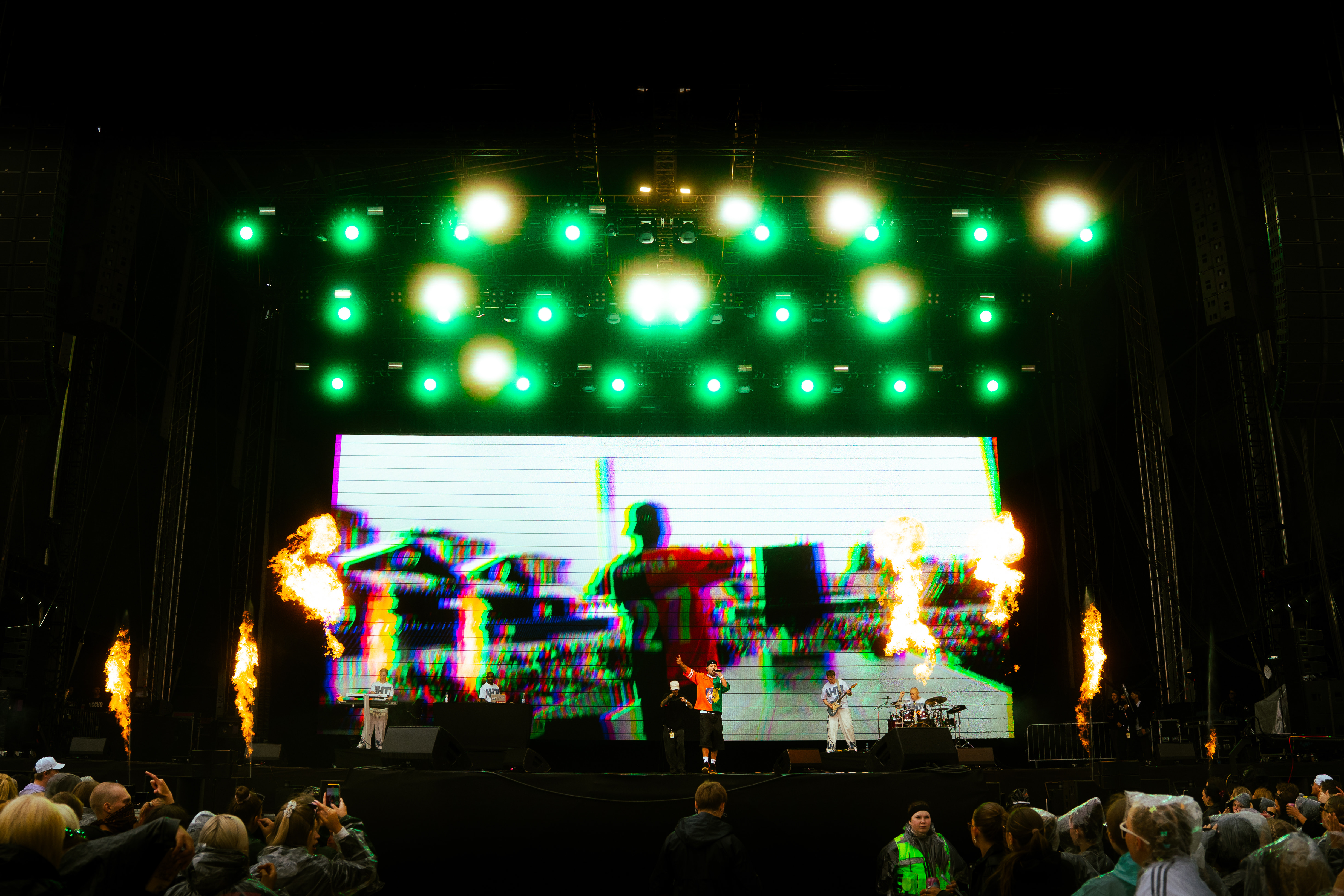 A live concert with performers on stage, green stage lights, fire effects, and a large colorful digital screen behind them, with an audience wearing rain ponchos.