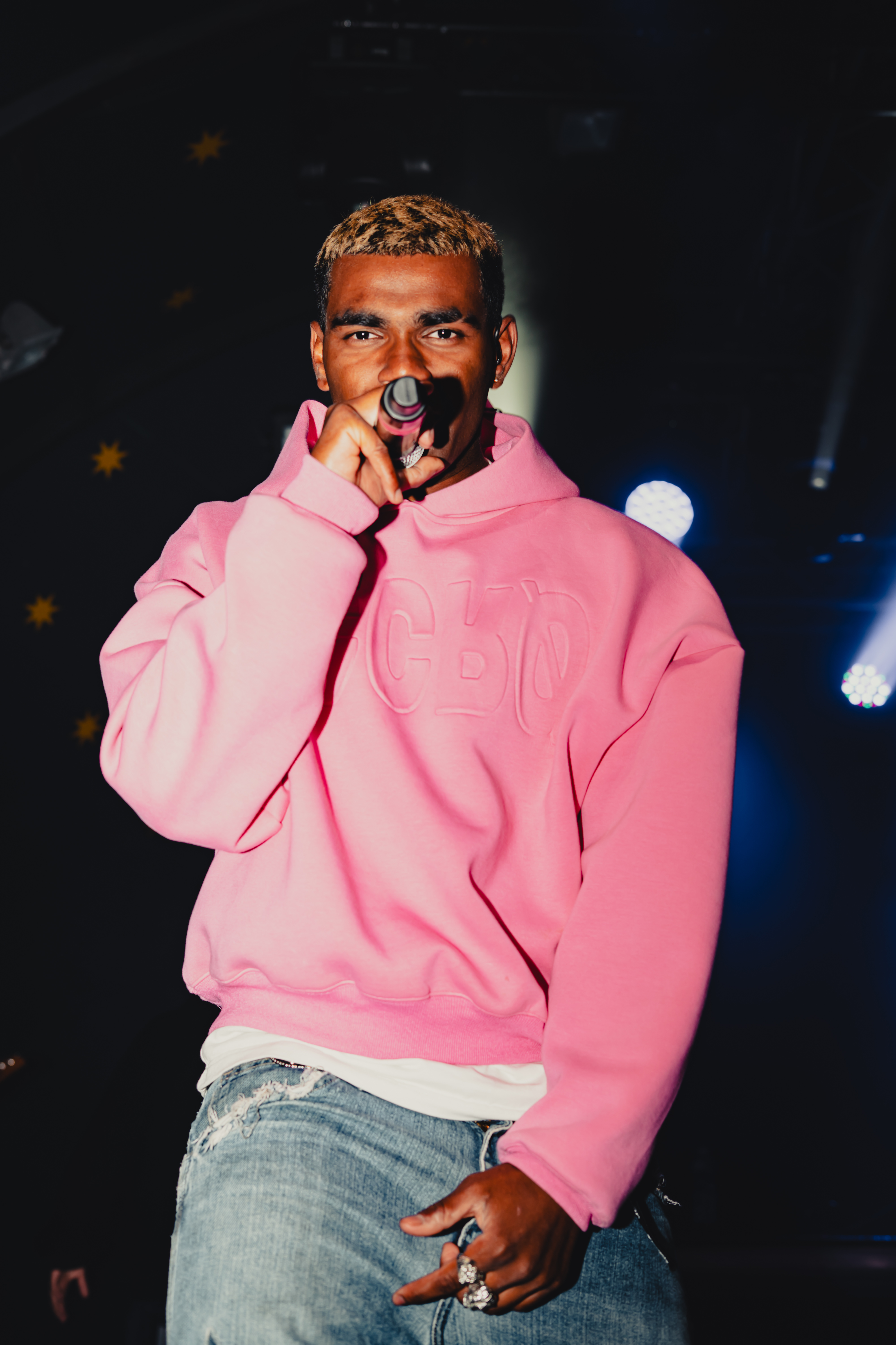 Man with blonde hair wearing a pink hoodie and ripped jeans performing on stage with a microphone.