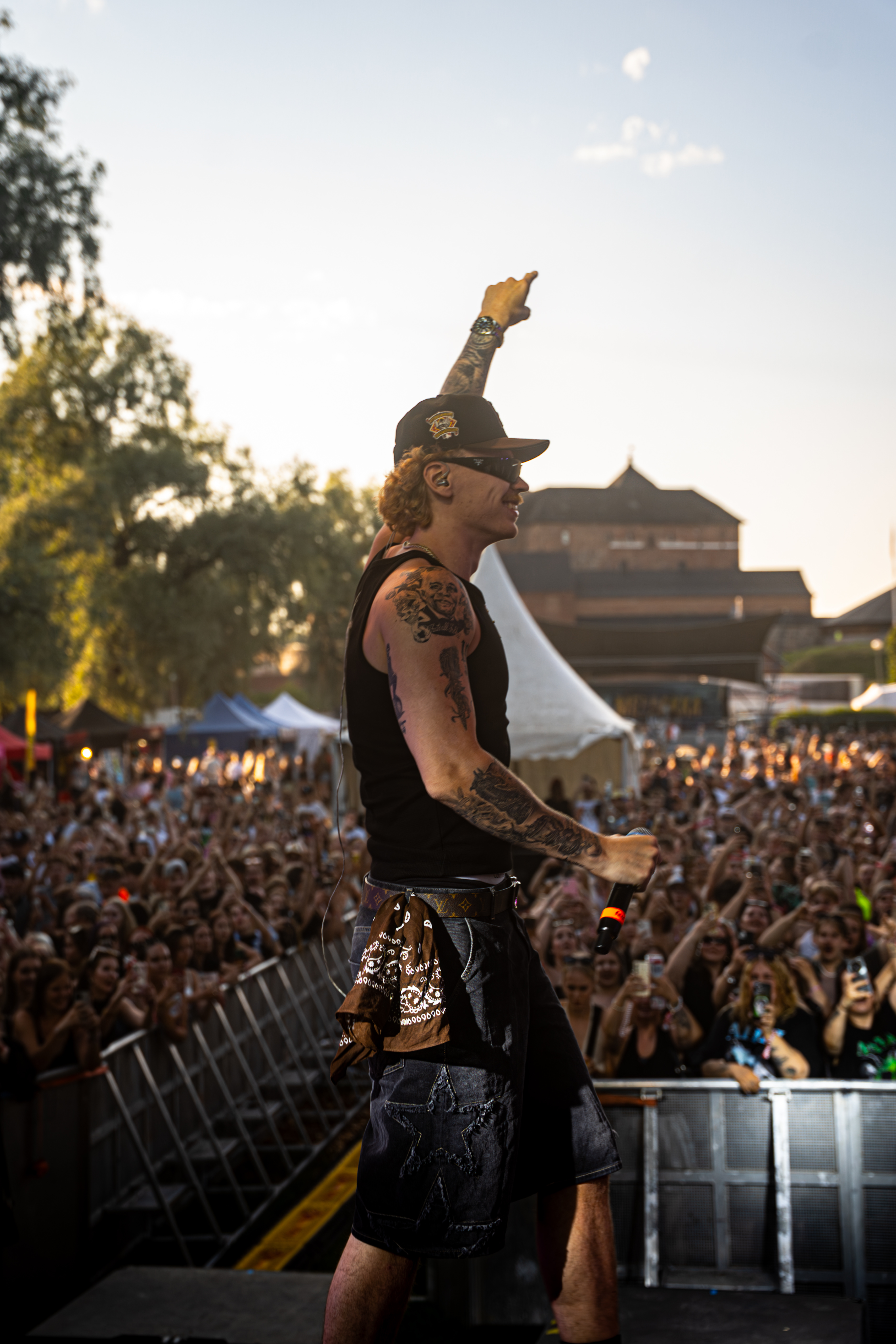 Tattooed male rapper in black tank top and cap performs on stage in front of a large outdoor crowd.