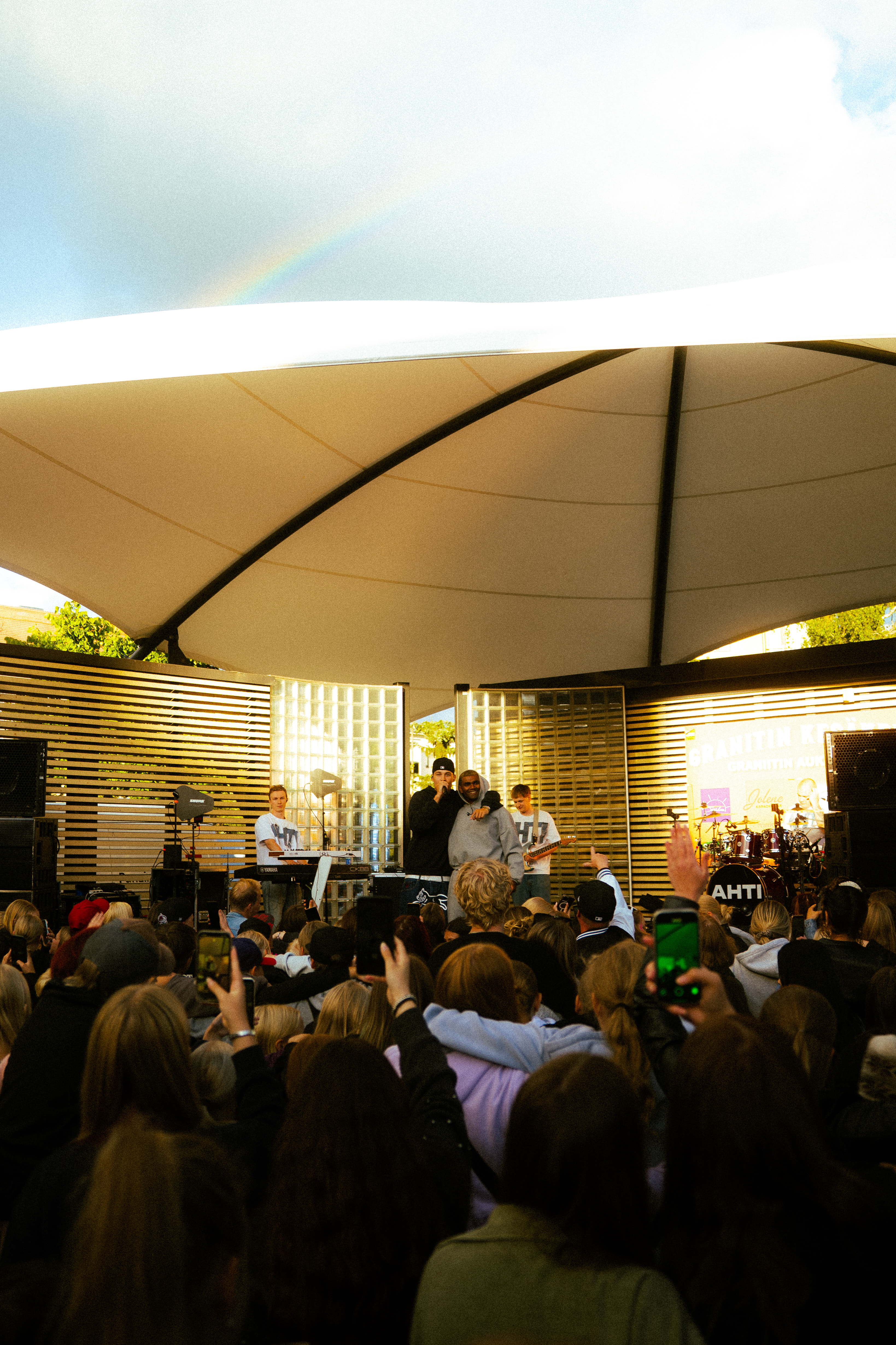 Crowd watching a live outdoor concert with two singers and musicians on stage under a large white canopy.