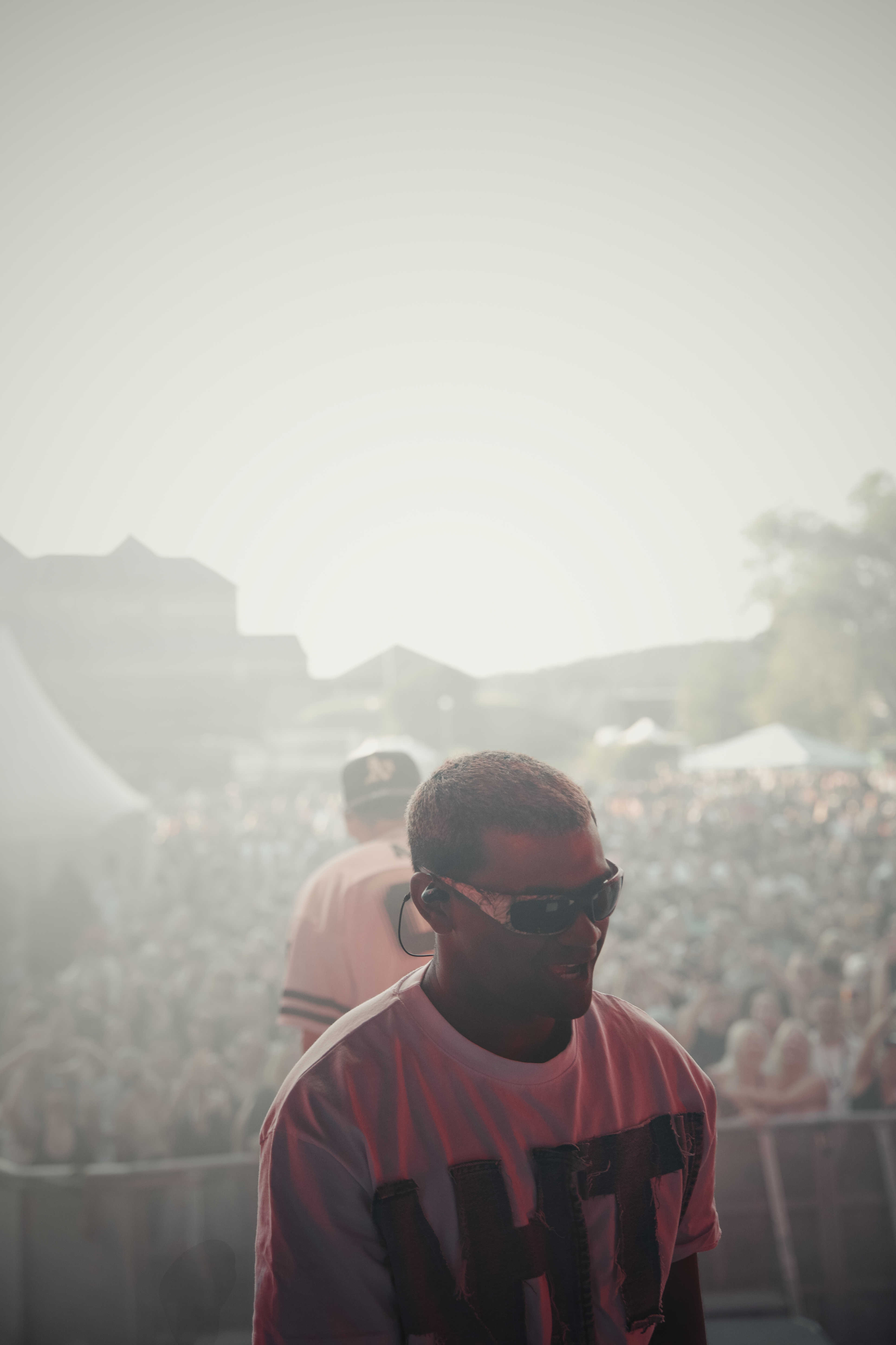 Man wearing sunglasses and a white shirt performing on stage with a large crowd blurred in the background.