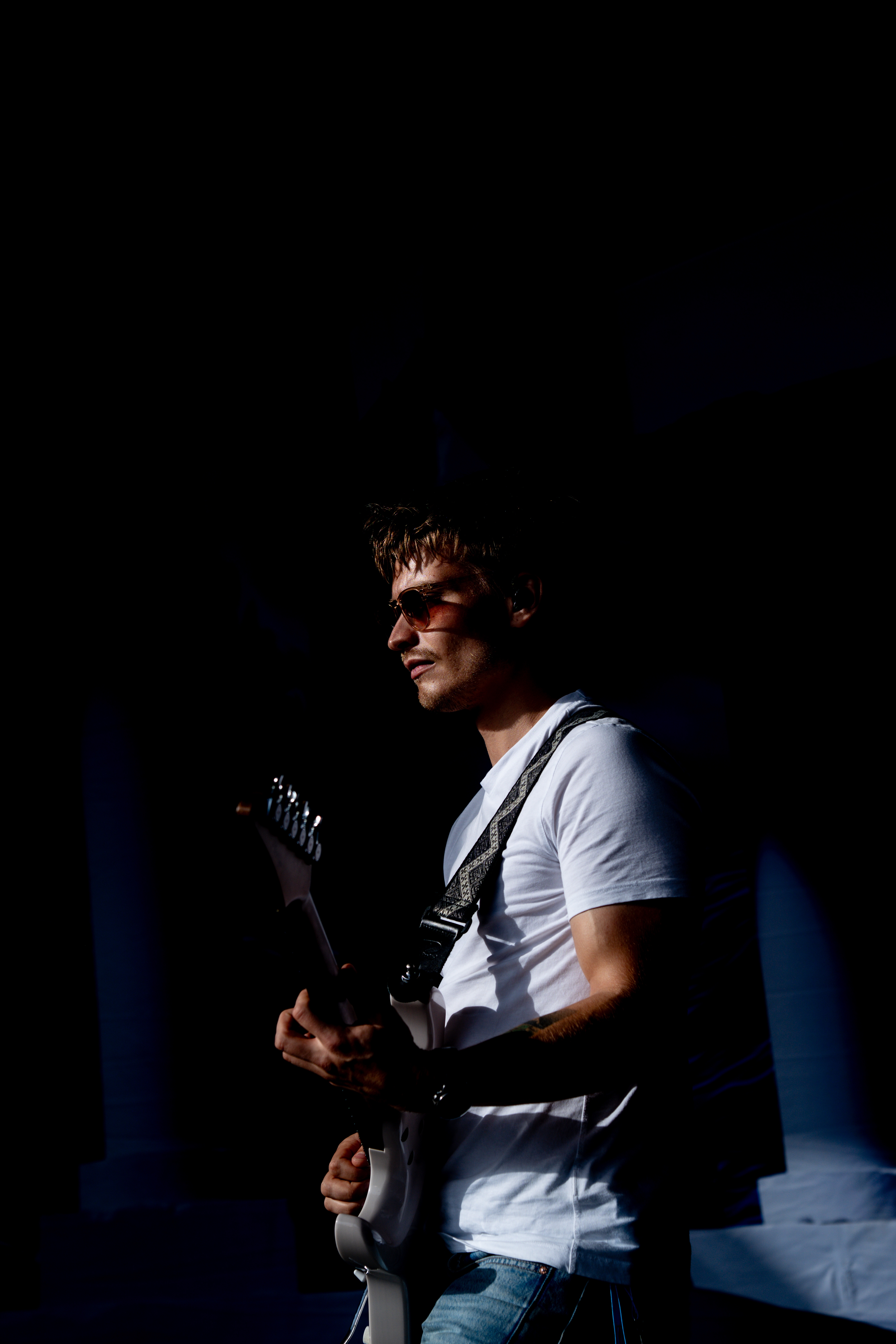 Man wearing sunglasses and a white t-shirt playing an electric guitar in low light.