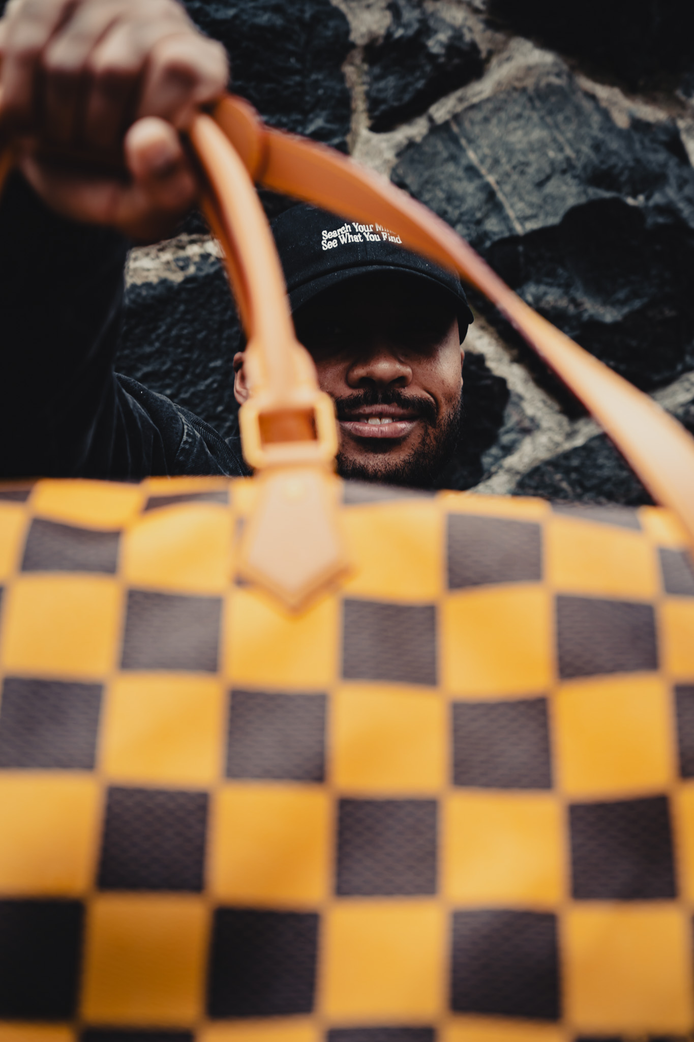 Man wearing a black cap with text 'Search Your Mind See What You Find' holding a yellow and brown checkered bag in front of a stone wall.