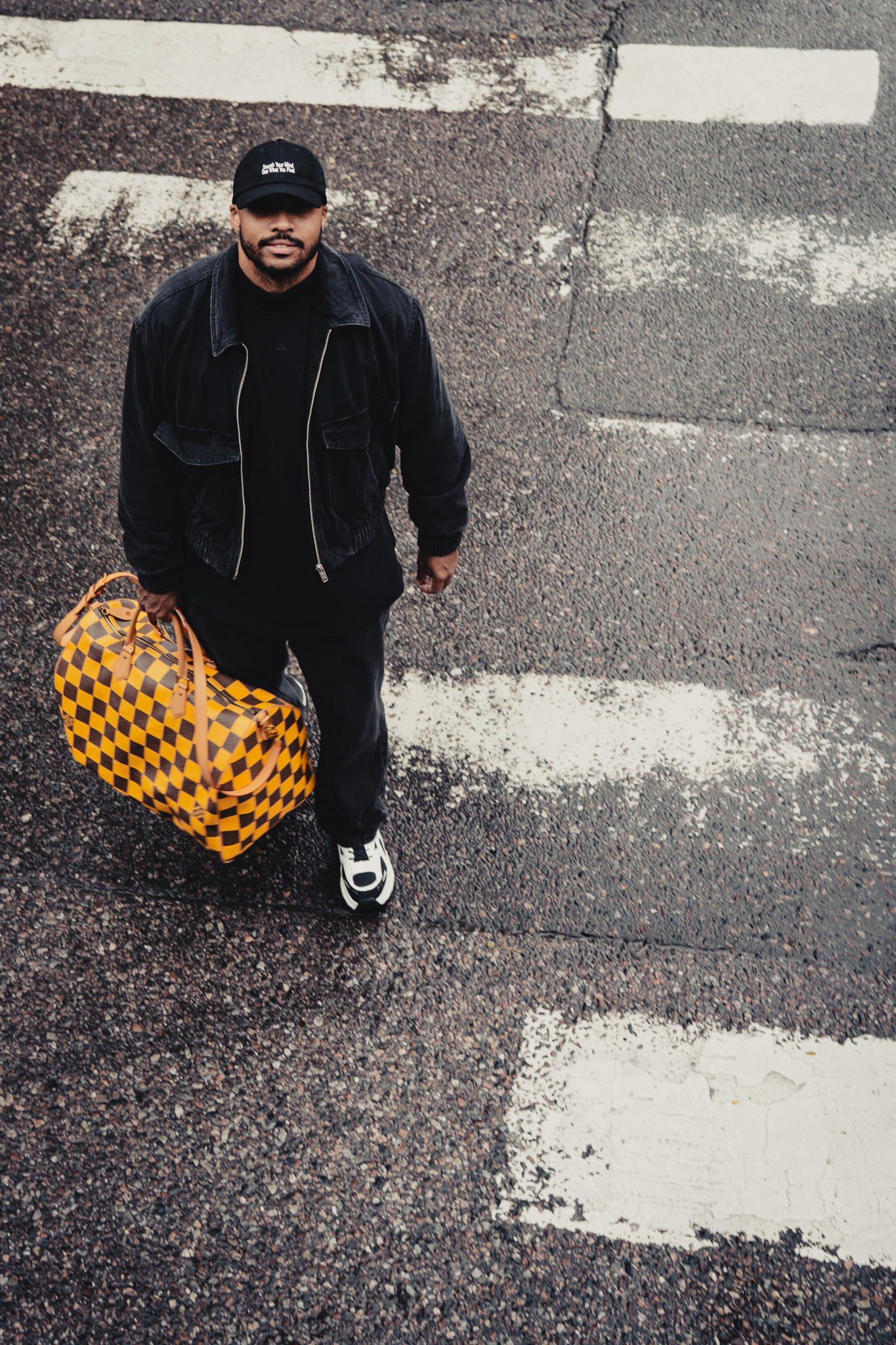 Man in black jacket and cap holding a yellow and brown checkered duffle bag standing on a textured street with faded white crosswalk markings.