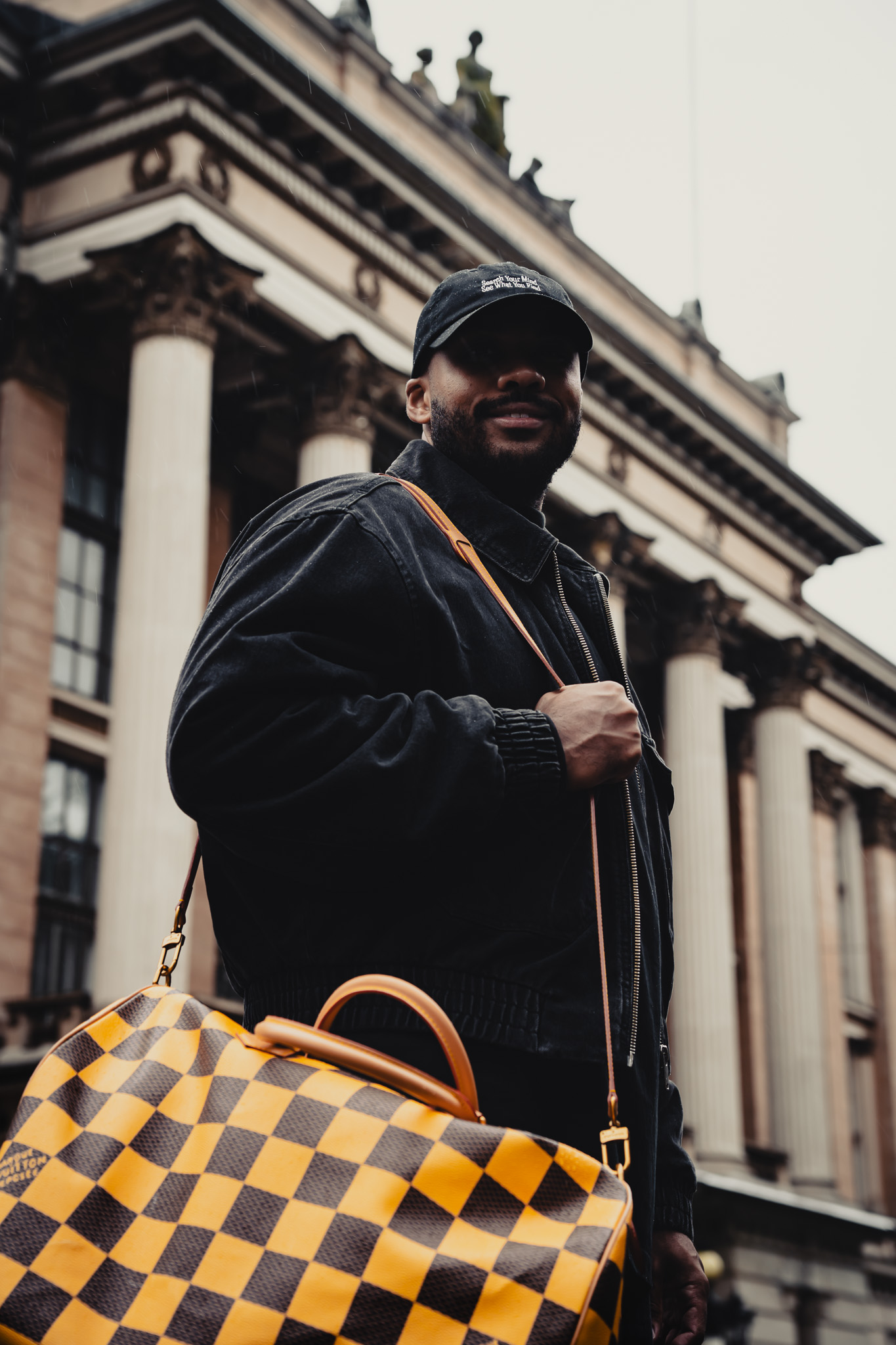 Man in black jacket and cap holding a yellow and brown checkered patterned bag in front of a building with tall columns.
