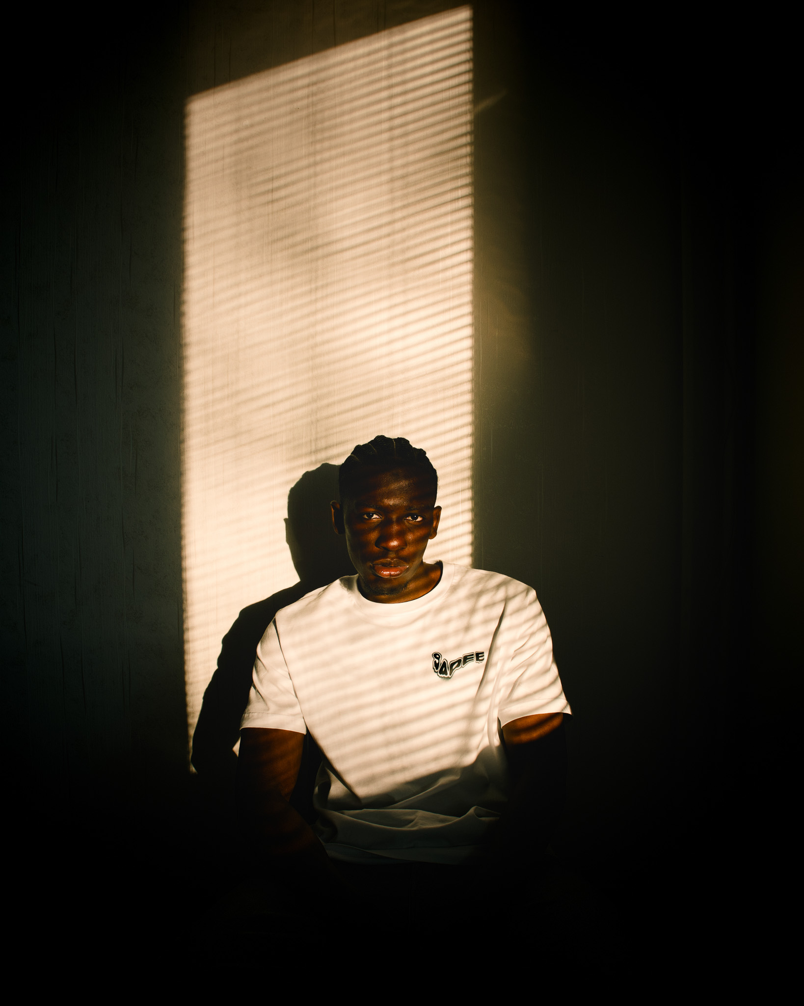 Man sitting in a dark room with striped light from window blinds casting shadows on his face and white t-shirt.