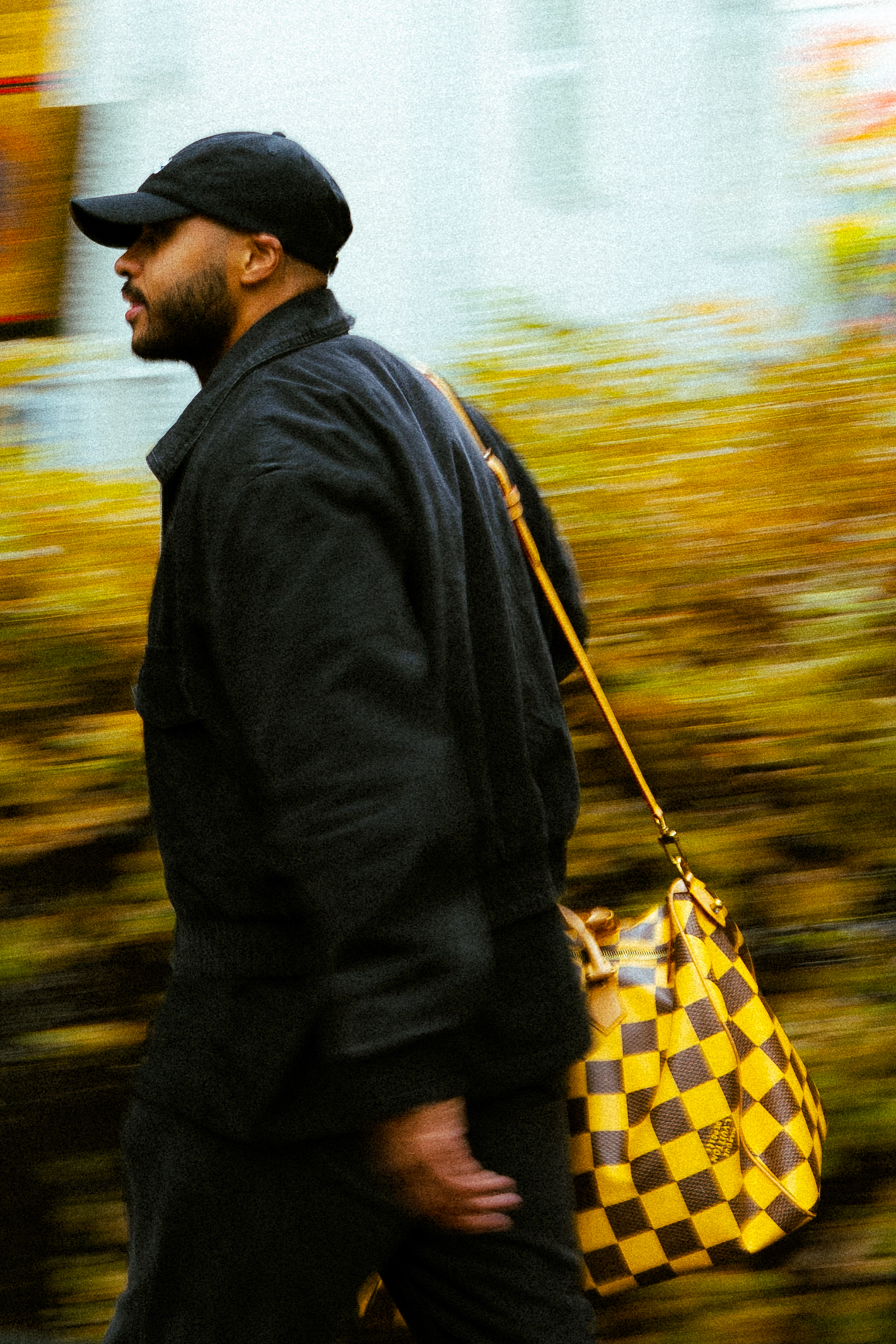 Man walking outdoors wearing a black jacket and cap with a yellow checkered handbag over his shoulder.