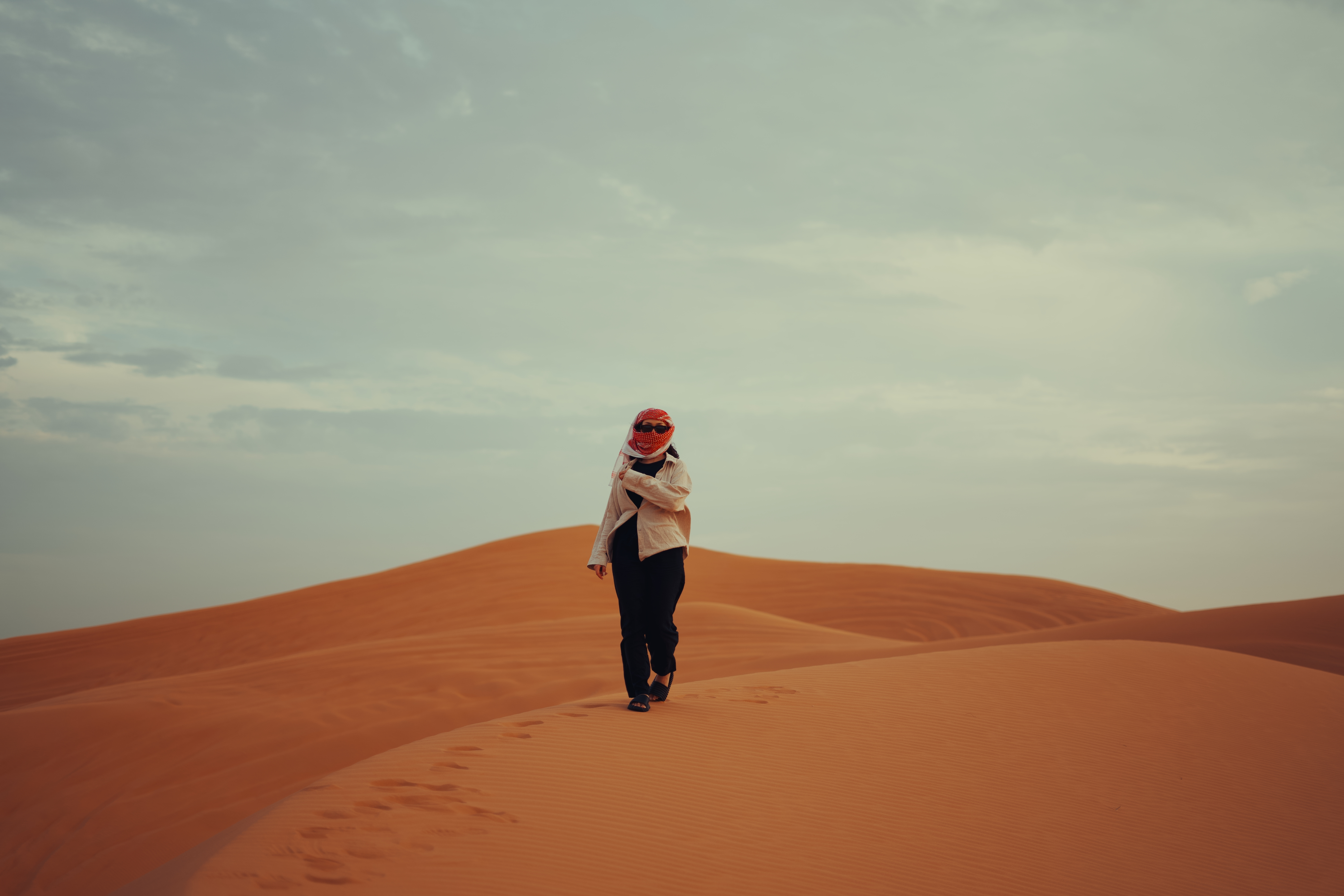Person wearing a light jacket, dark pants, and a red scarf walking alone on orange sand dunes under a cloudy sky.